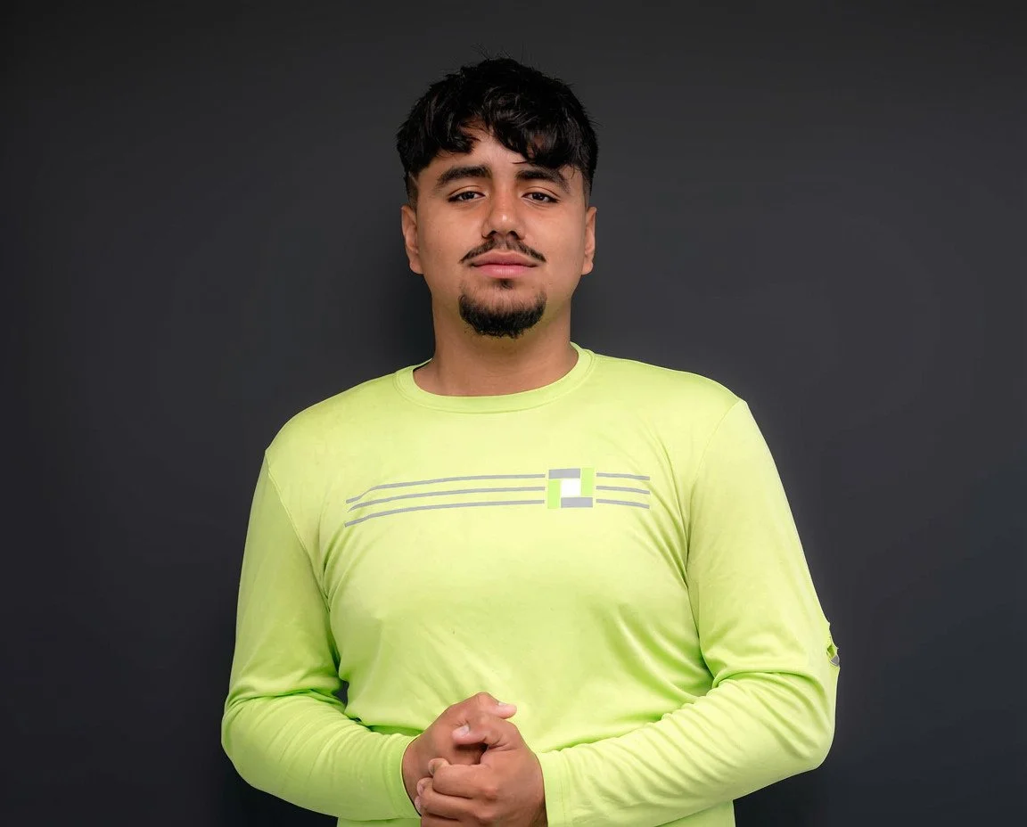 A young man with dark hair and a goatee wearing a bright yellow long-sleeve shirt stands in front of a dark gray background, looking directly at the camera with a slight smile.