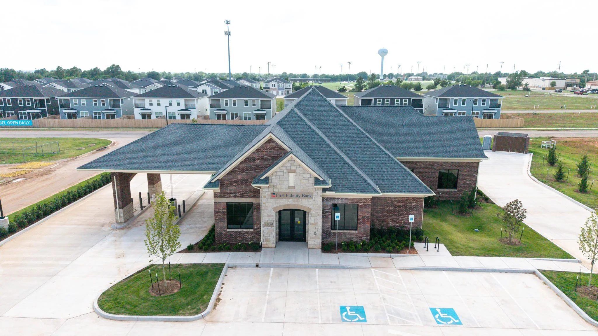 An aerial view of a commercial building with a brick facade, with a sign reading 'First Fidelity Bank.' There are parking spaces in front, including designated accessible spots, with a landscaped area and trees. In the background, there are residenti