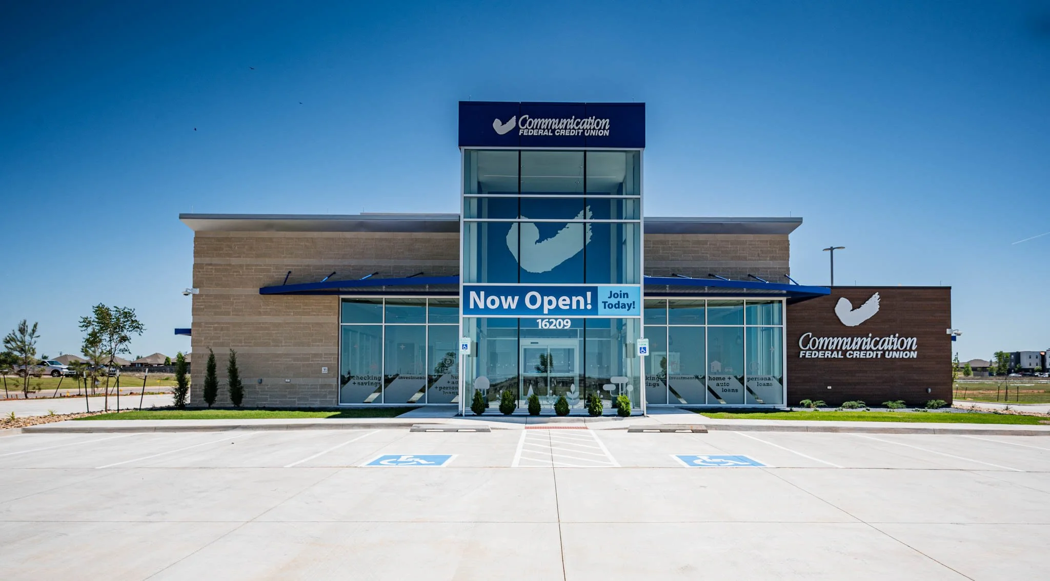 Newly opened Communication Federal Credit Union branch with a sign saying 'Now Open! Join Today!' located at 16209, with parking spaces, including designated handicap spots, and a clear blue sky.