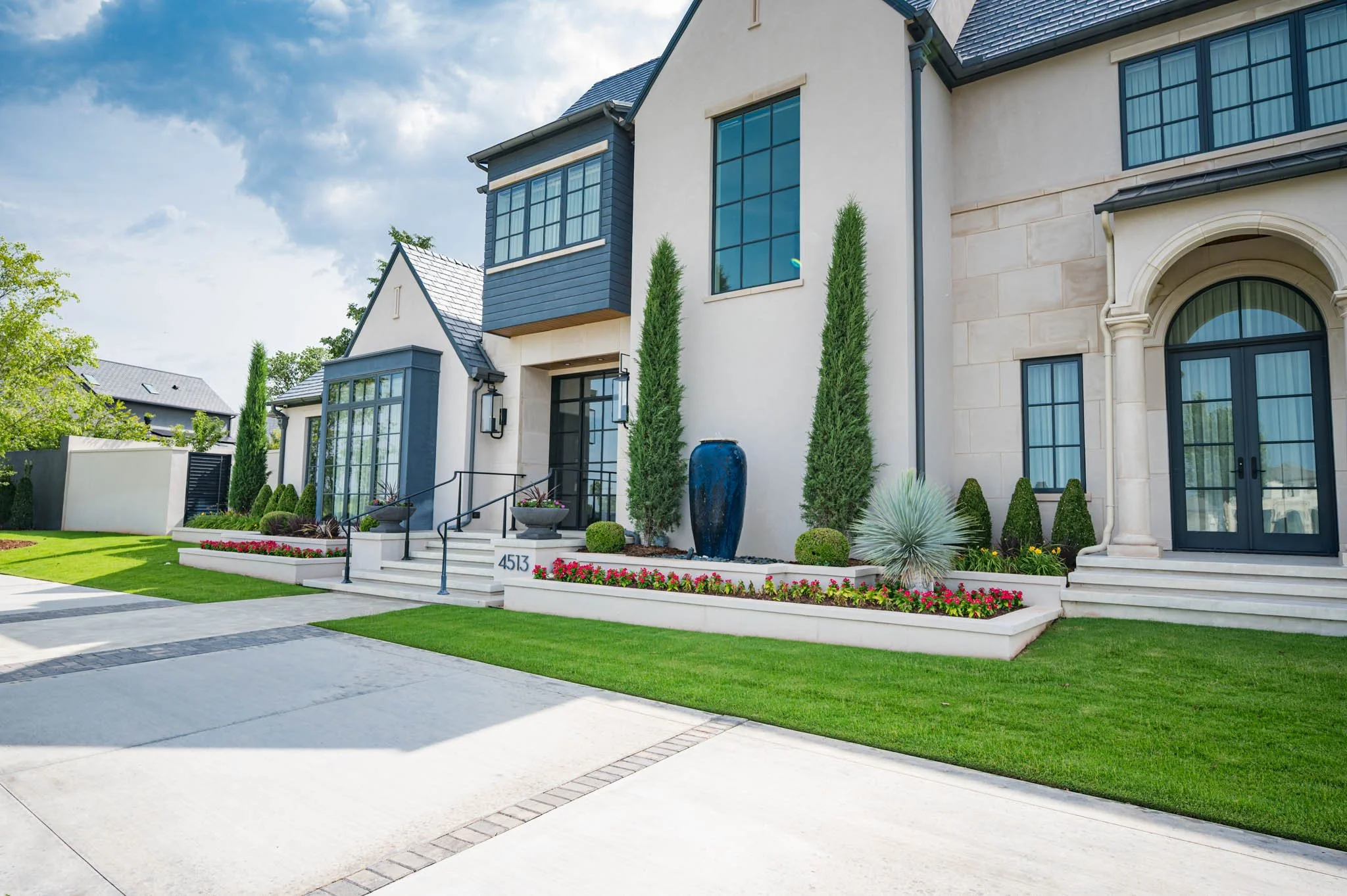 Modern house with large windows, surrounded by a well-maintained lawn and decorative plants, under a partly cloudy sky.