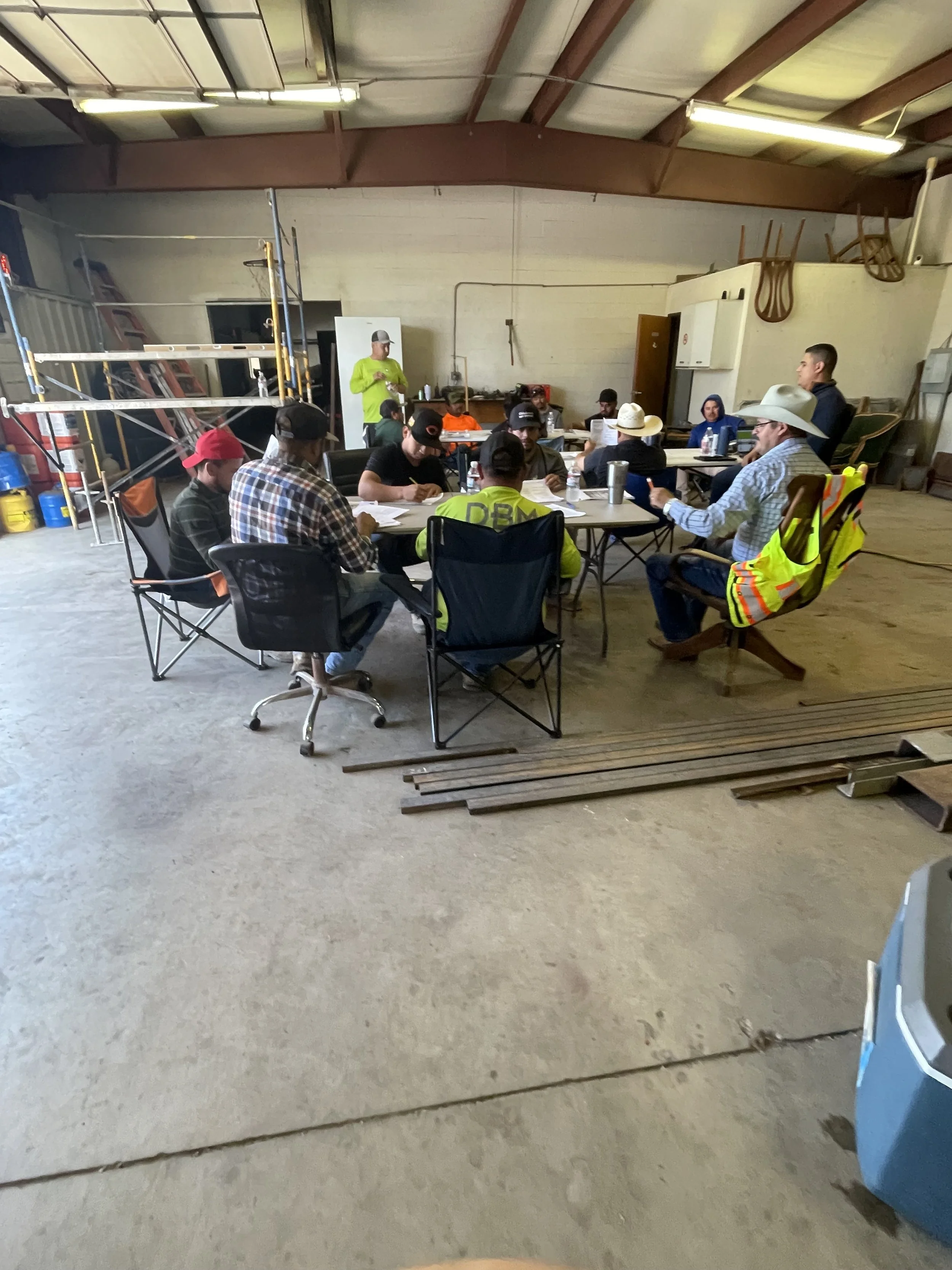 Meeting of several people wearing cowboy hats and casual work clothes sitting at tables in a warehouse or industrial space, with a person standing and speaking.