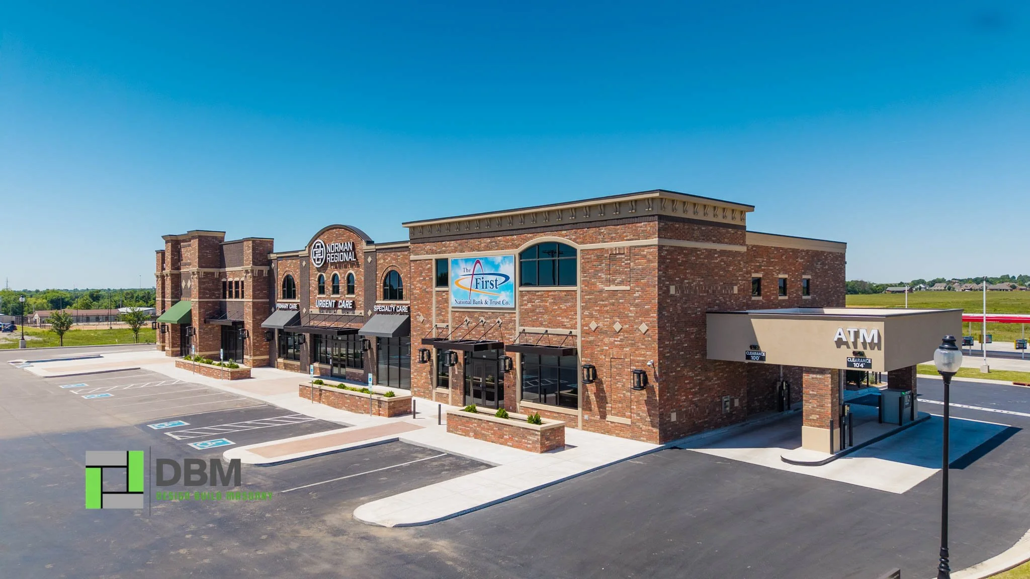 A brick commercial building with signs for urgent care, specialty care, Norman Regional, and The First National Bank & Trust. There is an ATM on the side and a parking lot in front with several parking spaces, including accessible ones. The sky is clear and blue.