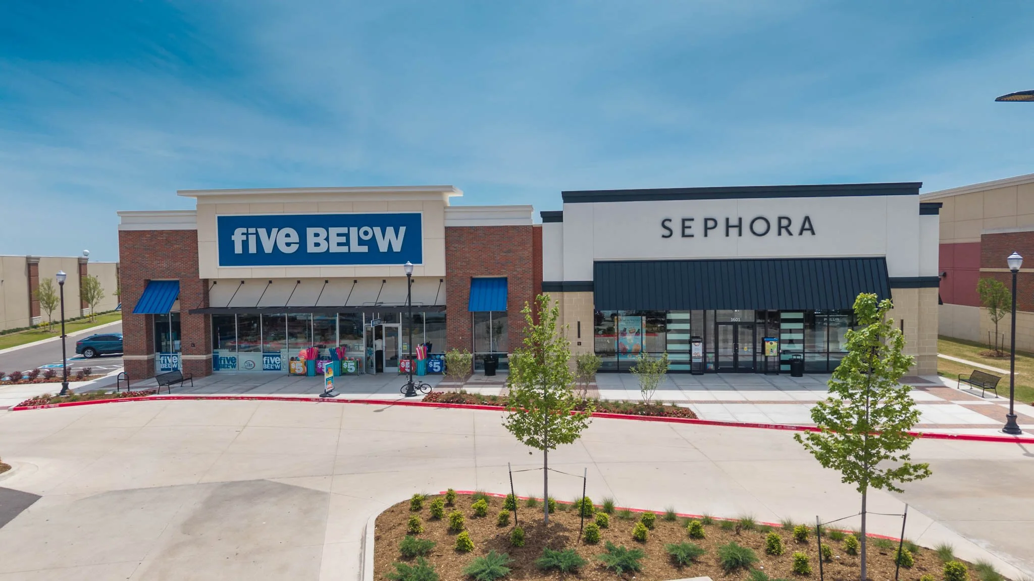 A shopping plaza with Five Below and Sephora stores, clear blue sky, trees, lamp posts, parked car, and a bicycle