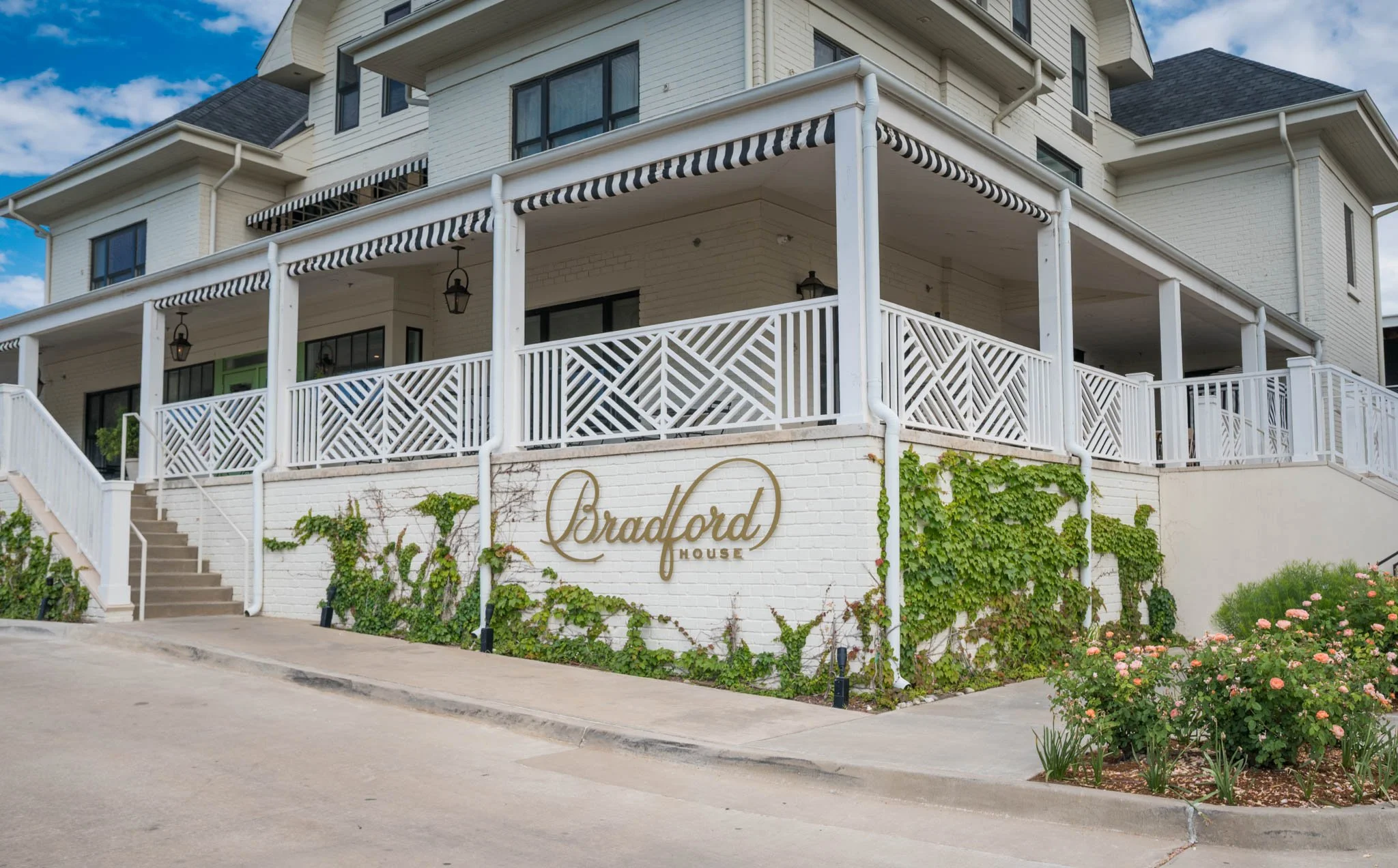 A white multi-story building with a large porch and black and white striped retractable awnings. There are black lantern-style lights hanging from the ceiling of the porch. The building has a sign that reads 'Bradford House' on the white brick wall n