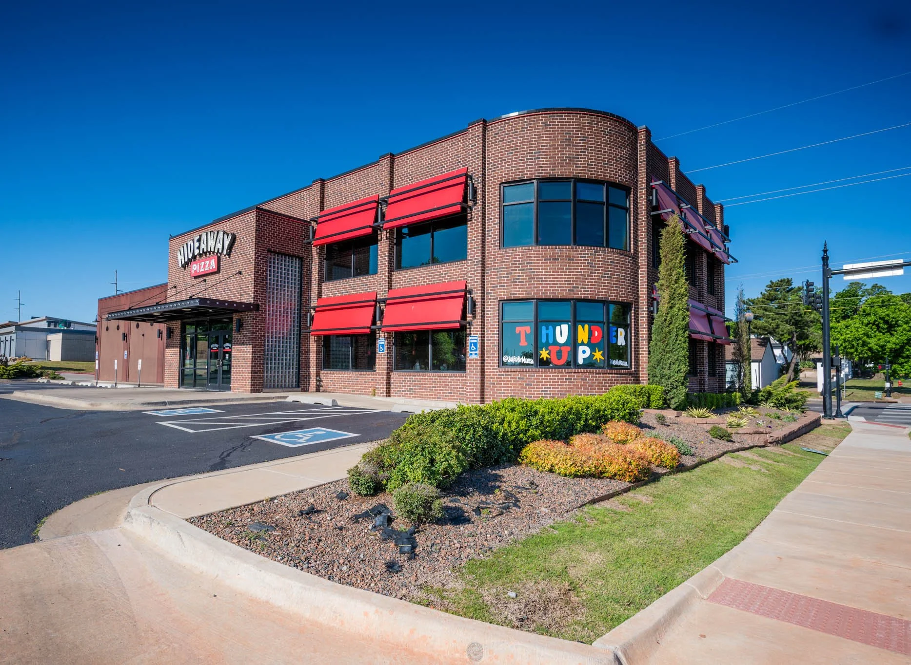 A two-story brick building with red awnings and large windows, with a sign that reads "Hideaway Pizza." The parking lot in front has designated handicapped spaces, and there is a garden bed with bushes and plants along the sidewalk.
