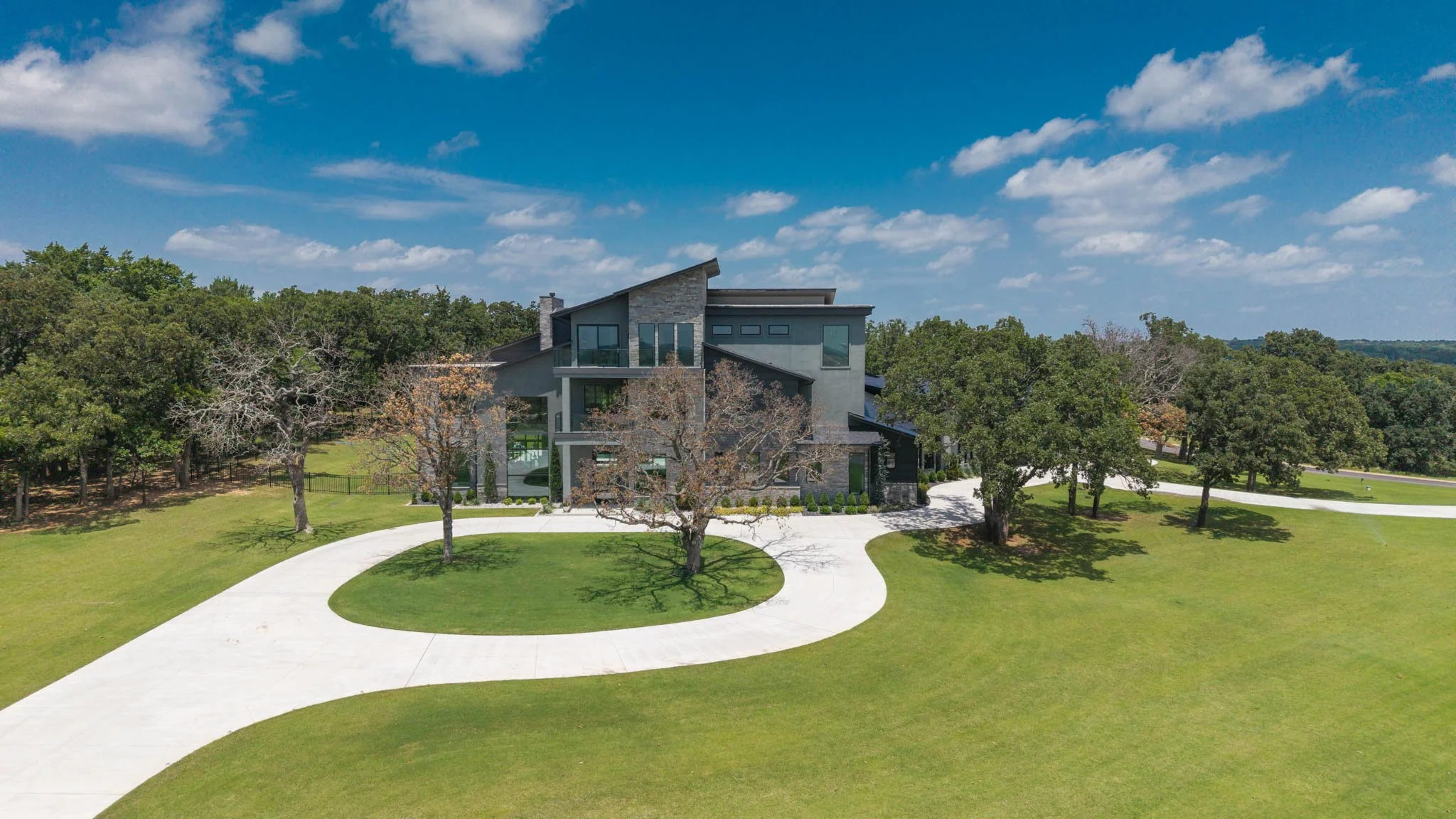 Modern house surrounded by lawn and trees, with a curved driveway leading to the entrance under a partly cloudy blue sky.