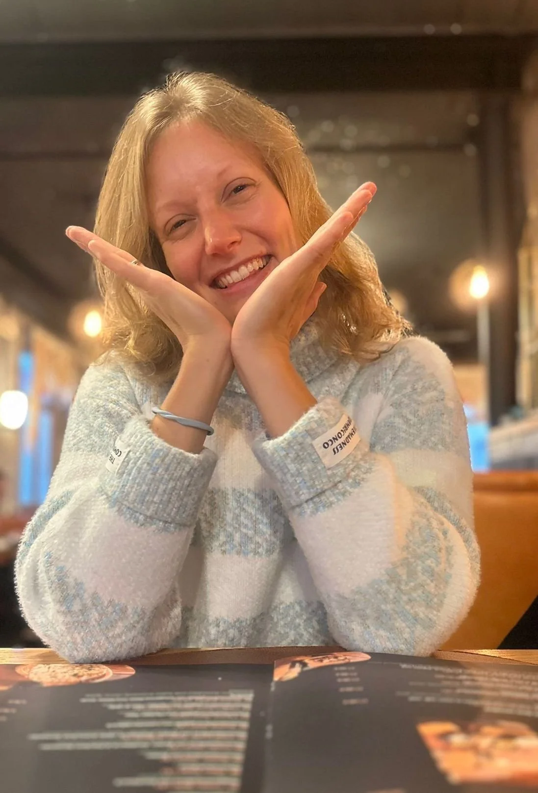 A blond, fair-skinned woman smiling, resting her face in her hands, sitting at a table in a cozy, warmly lit restaurant.