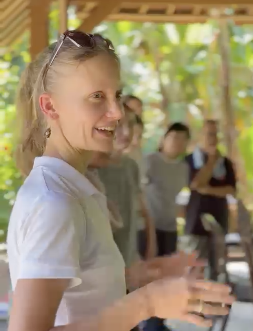 A blonde, fair-skinned woman wearing a white shirt and sunglasses, smiling and talking to a group of people in an outdoor setting with greenery.