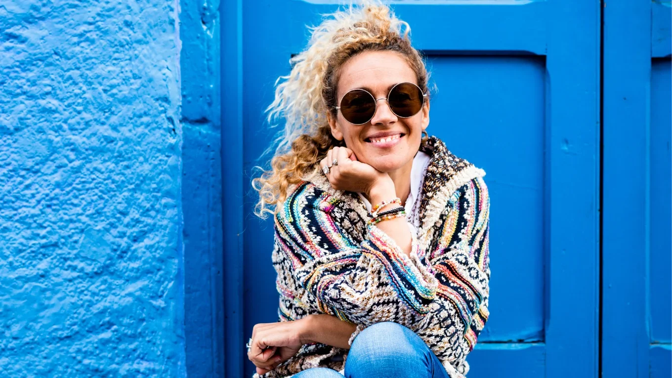 A woman with curly blonde hair wearing sunglasses, a colorful patterned sweater, and light blue jeans, sitting against a bright blue wall and smiling at the camera.