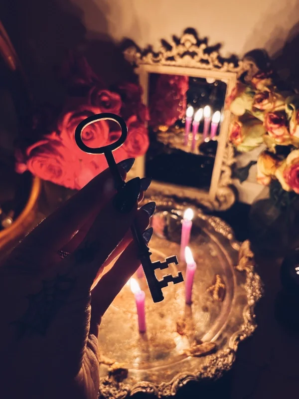 A person's hand holding an antique skeleton key in front of a vintage mirror and a candelabra with pink candles, surrounded by flowers, creating a dim, decorative scene.