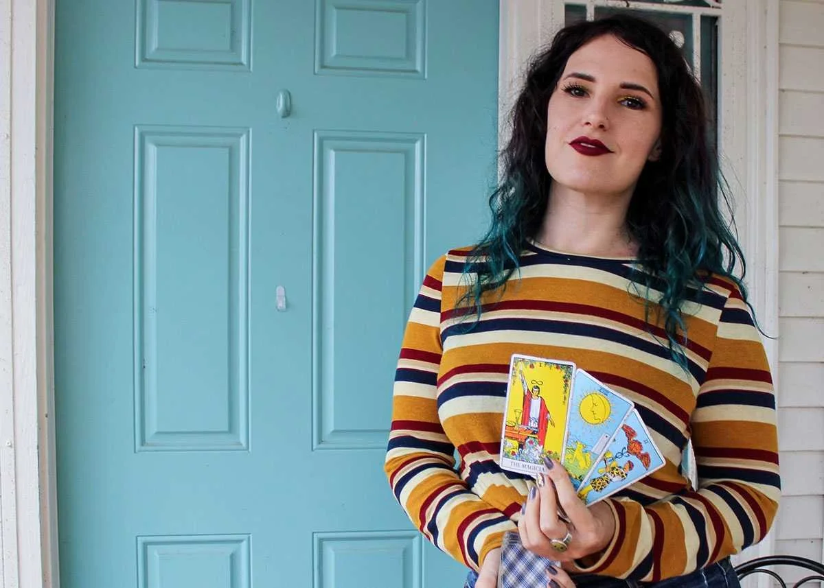 A woman with dark wavy hair with blue tips, wearing a striped sweater in mustard yellow, navy, red, and white, holds a few tarot cards in front of her. She stands outside next to a light blue door on a house with white siding.