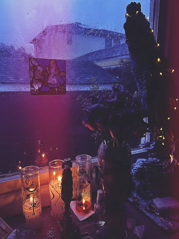 A rainy window with a house in the background, a stained glass angel decoration, lit candles, and a flower arrangement on a windowsill.