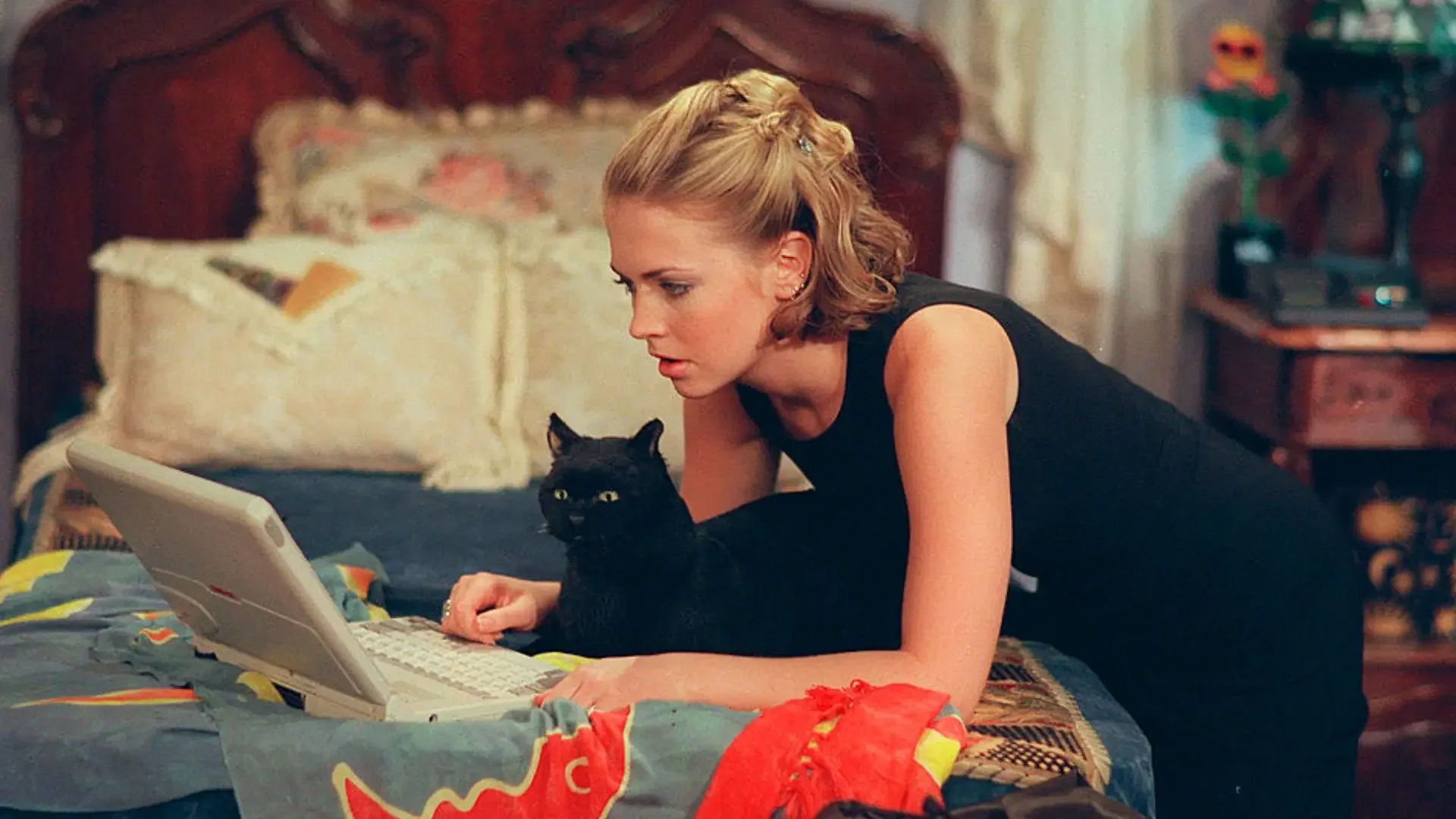 A woman with short blonde hair and a black sleeveless top lying on a bed, using a laptop. Next to her is a black cat with yellow eyes, also on the bed, looking at the laptop. The bed has colorful blankets and pillows and the background includes a wooden headboard and a nightstand with decorations.