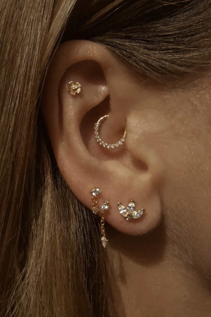 Close-up of a person's ear with multiple earrings, including a flower-shaped stud, a diamond hoop, a dangling earring with small diamonds, and a leaf-shaped cluster of gemstones. The person has light brown hair.