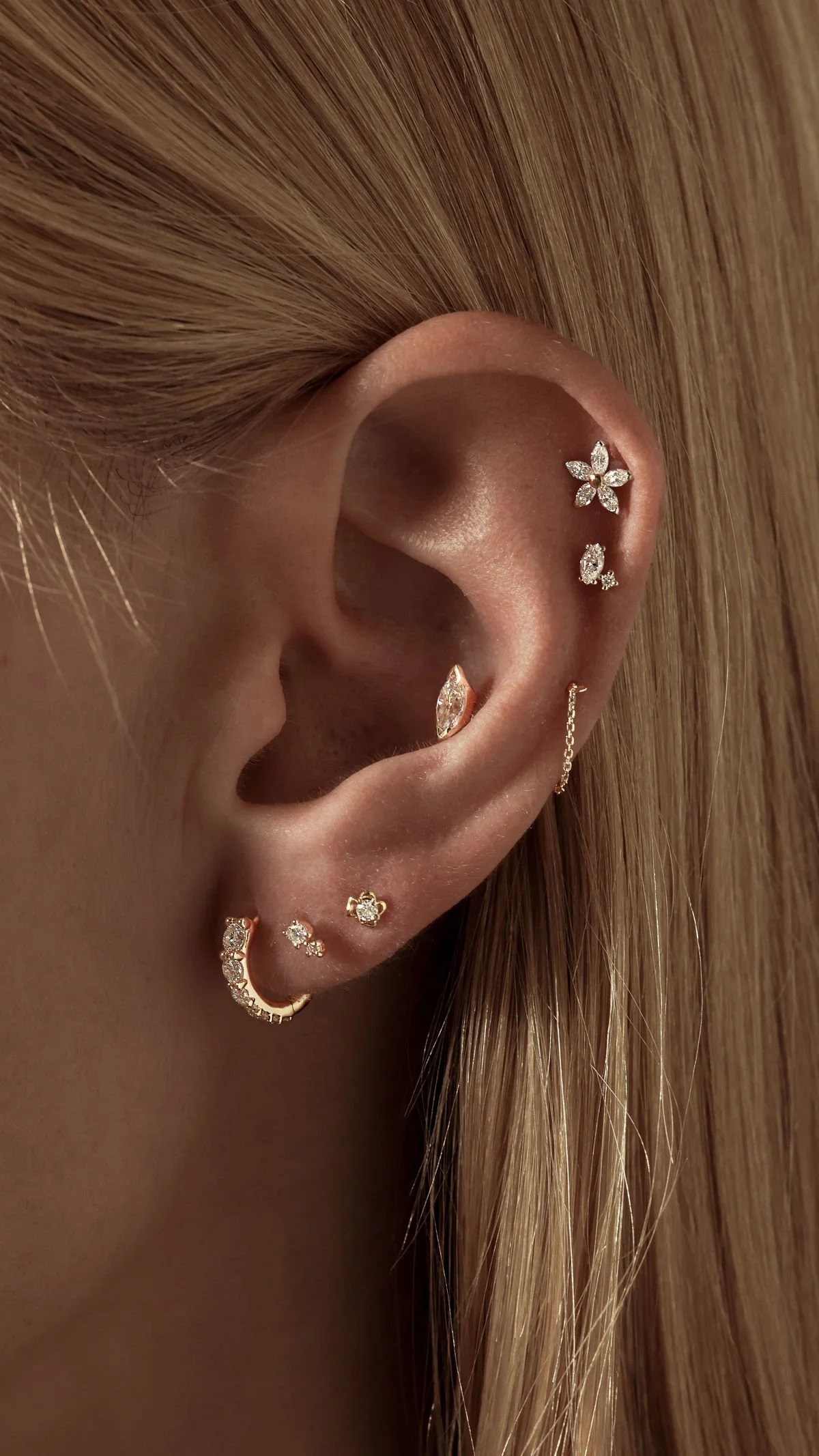 A close-up of a woman's ear decorated with multiple gold and diamond earrings, including a floral-shaped ear cuff, a chain-connected earring, and small stud earrings, with blonde hair partially visible.