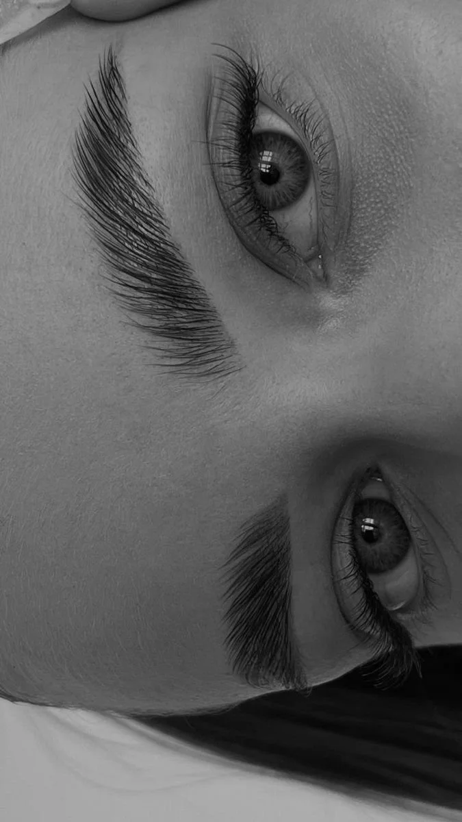 Close-up black and white photo of a person's face reflected upside down in water, focusing on the eye, eyebrows, and eyelashes.