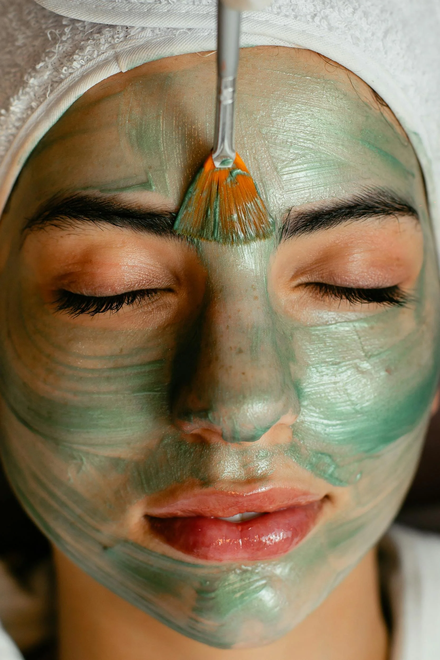 Close-up of a woman with green facial mask being applied with a brush on her forehead.