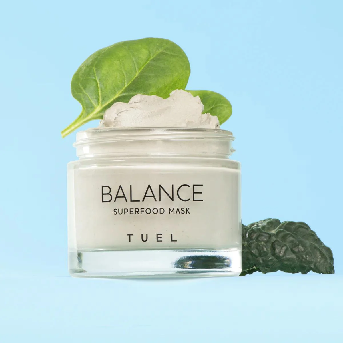 A jar of Balance Superfood Mask with whipped cream-like content topped with green leaves and rested against a blue background.