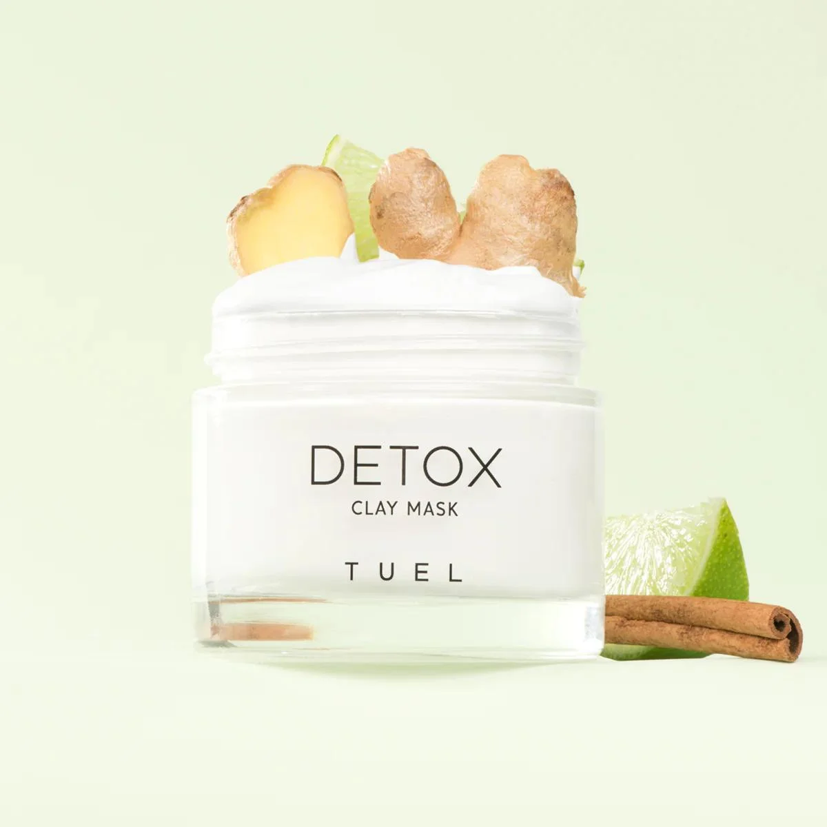 A jar of detox clay mask labeled 'TUEL' with sliced lime, ginger, and cinnamon sticks around it on a light green background.