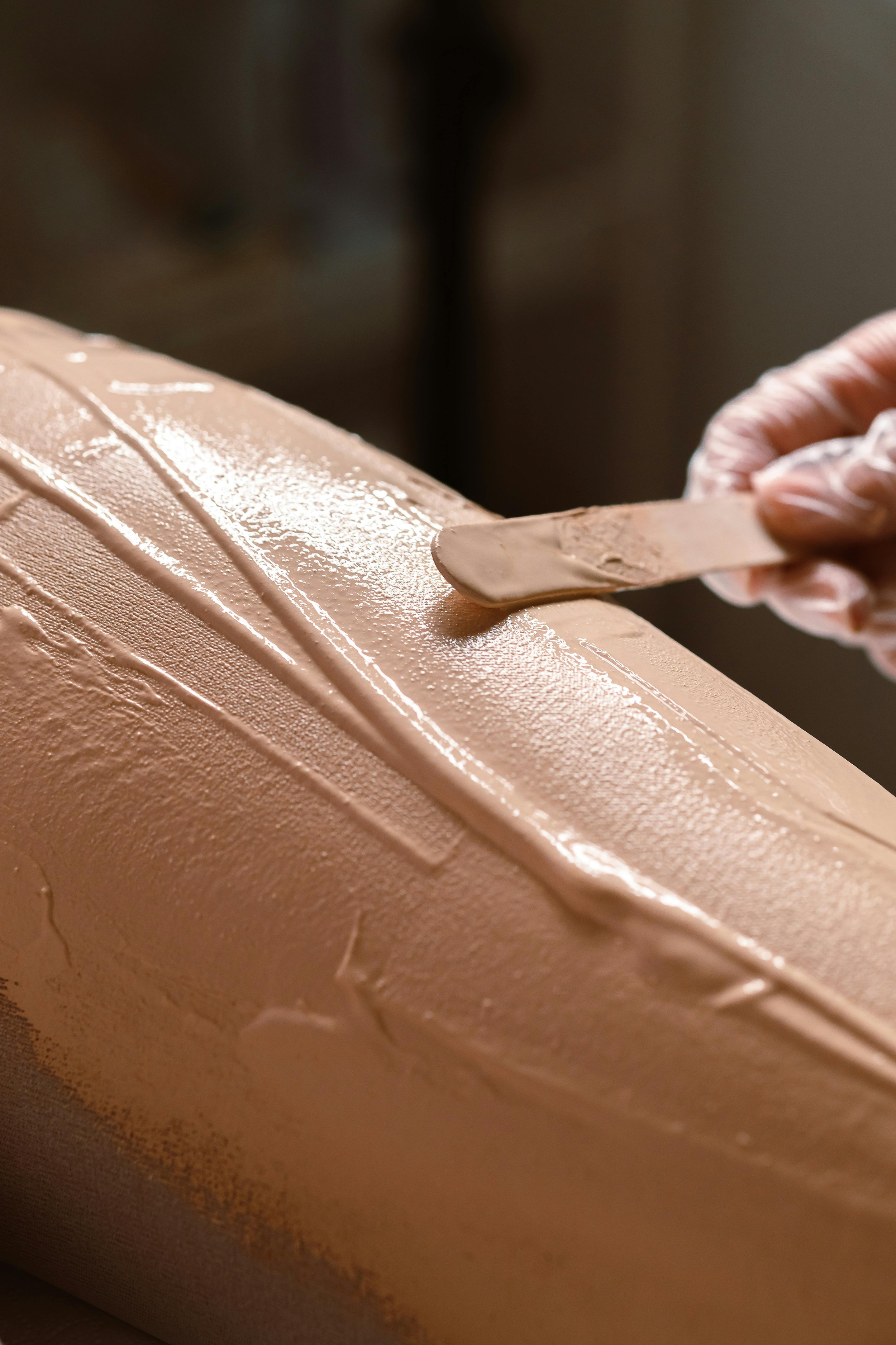 Close-up of a hand spreading beige cream or frosting onto a cake with a spatula in a kitchen or bakery setting.