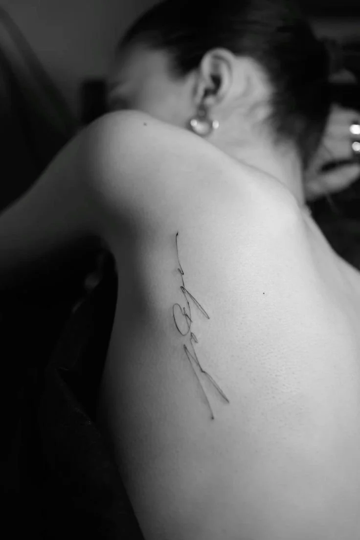 Black and white photo of a person with short hair, showing their back and shoulder, with a tattoo of the word 'love' in cursive on their upper back.