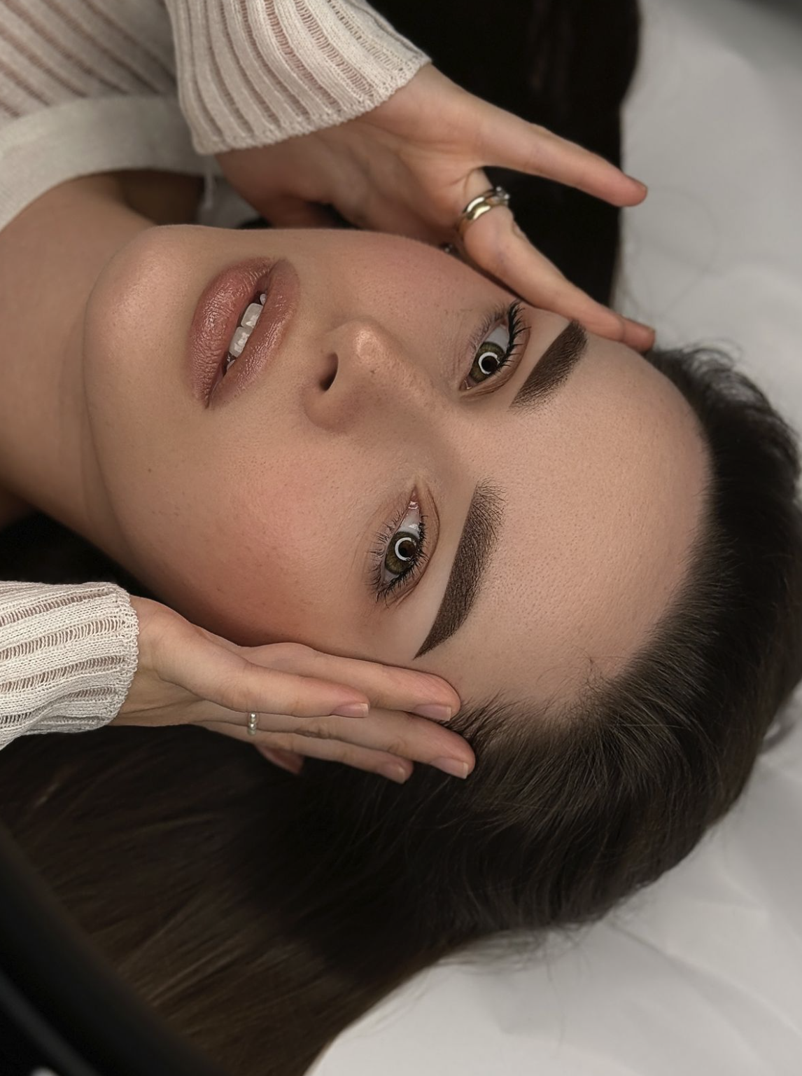 Close-up of a woman lying down, with her eyes open, showcasing her well-groomed eyebrows and makeup, while someone gently holds her face and head.