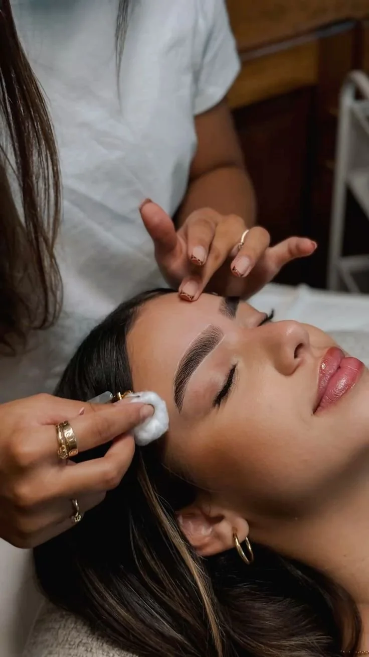 A woman receiving a facial treatment while lying down with closed eyes, with a person cleaning her face using a cotton pad.