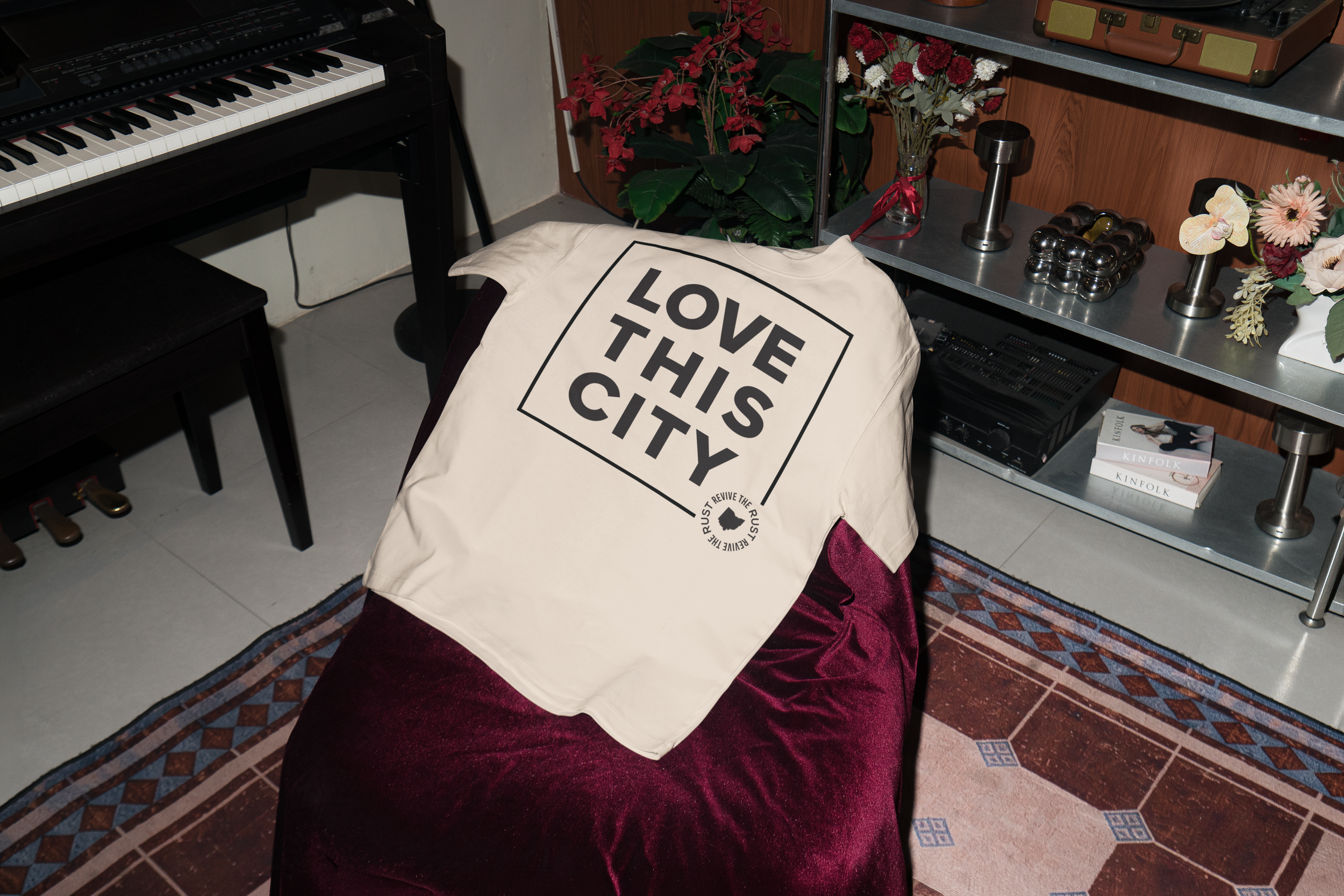 A beige tote bag with the words "LOVE THIS CITY" in black capital letters, placed on a maroon velvet surface in a room with a patterned carpet and a metal shelf holding flowers, a lamp, and a vintage radio, next to a keyboard and a small black speaker.