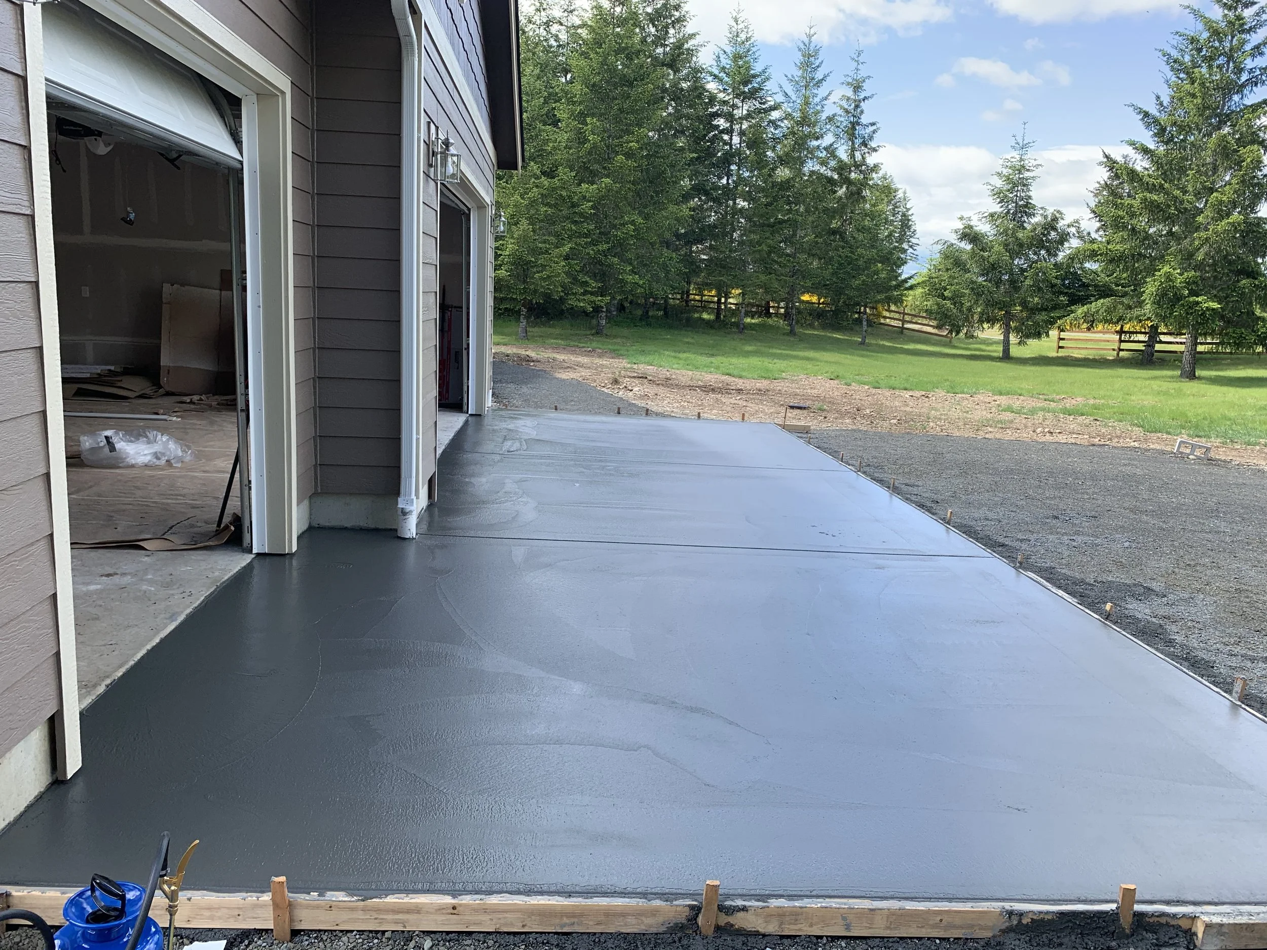 Newly poured concrete slab in a backyard with a house on the left and a line of trees in the background.