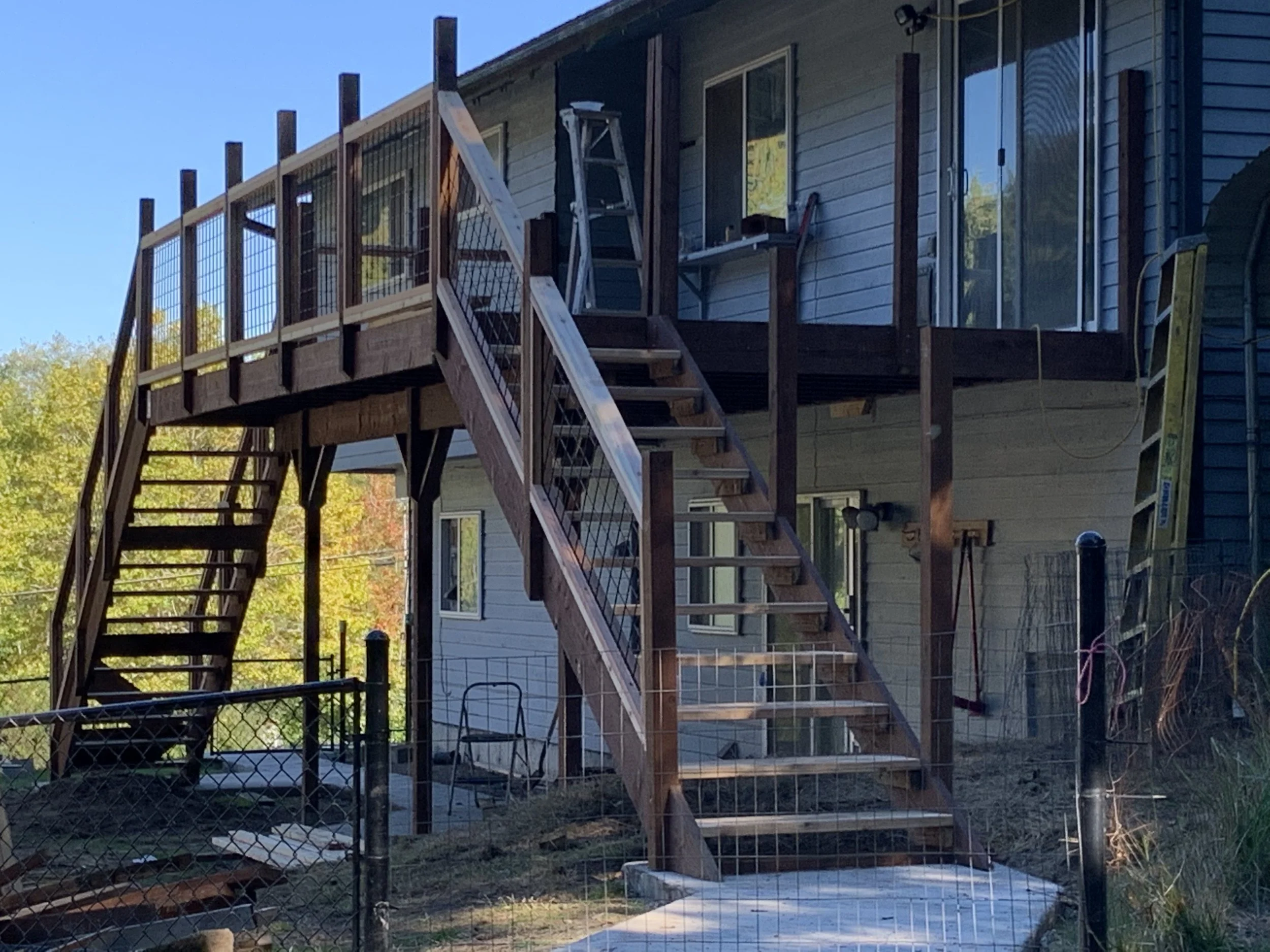 Backyard under construction with wooden deck and stairs, a ladder resting against the house, and a black chain-link fence surrounding the area.