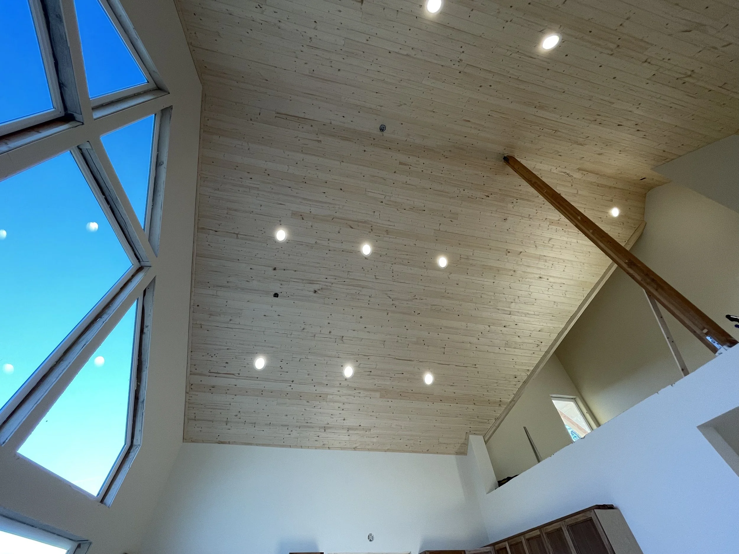 Interior view of a room with a sloped wooden ceiling, ceiling lights, and large windows showing a blue sky.