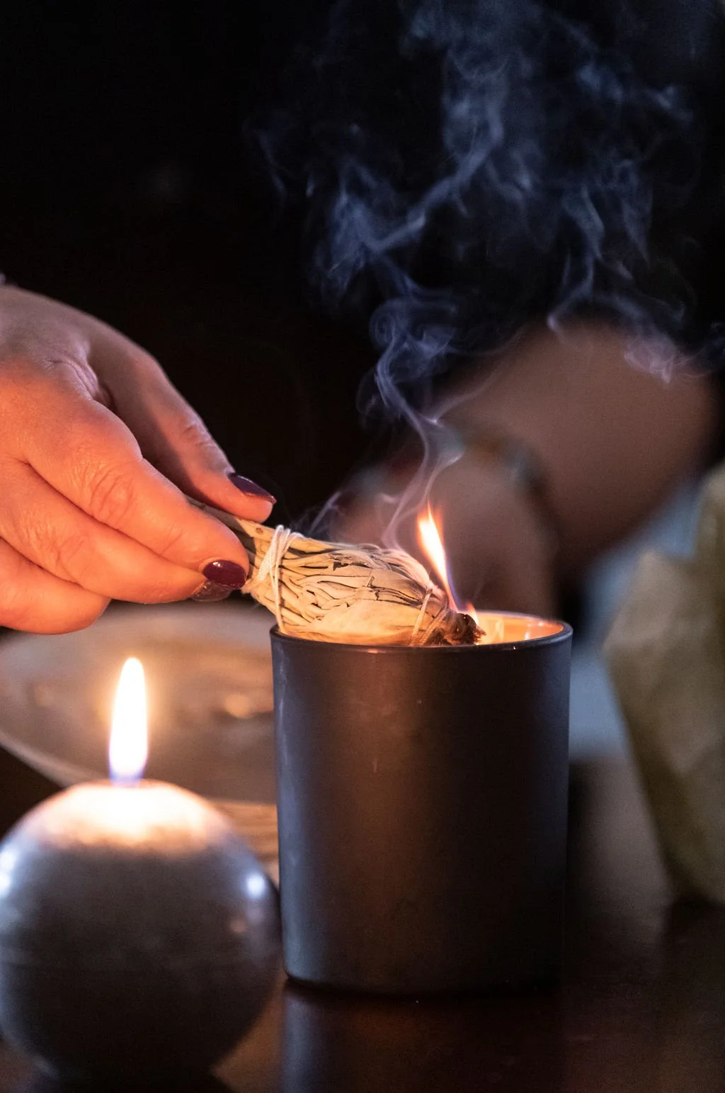 A person is lighting a sage bundle with a match near candles, with smoke rising in a dimly lit setting.