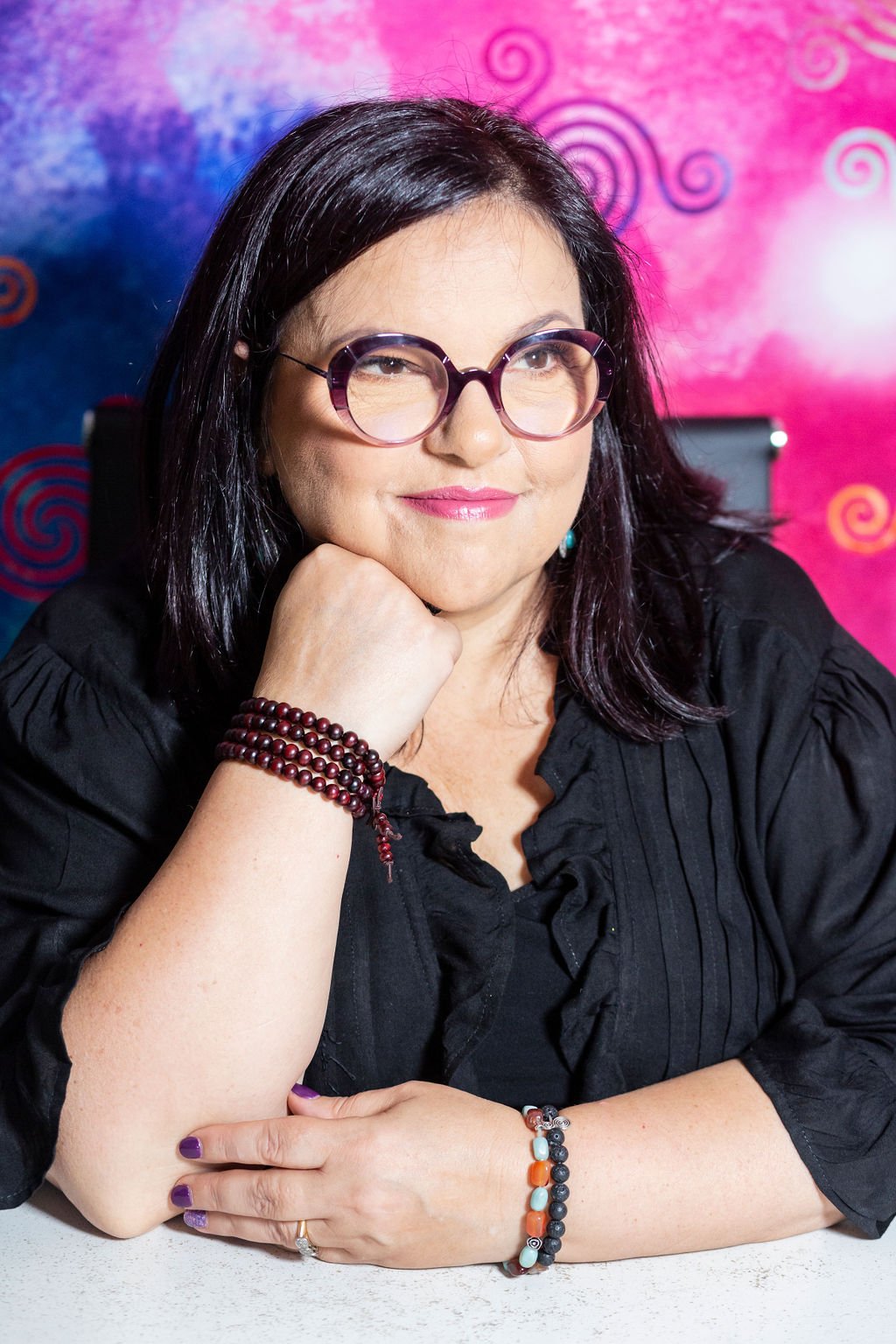 A woman with black hair, wearing glasses, jewelry, and a black blouse, sitting with her chin resting on her hand, in front of a colorful, abstract background.