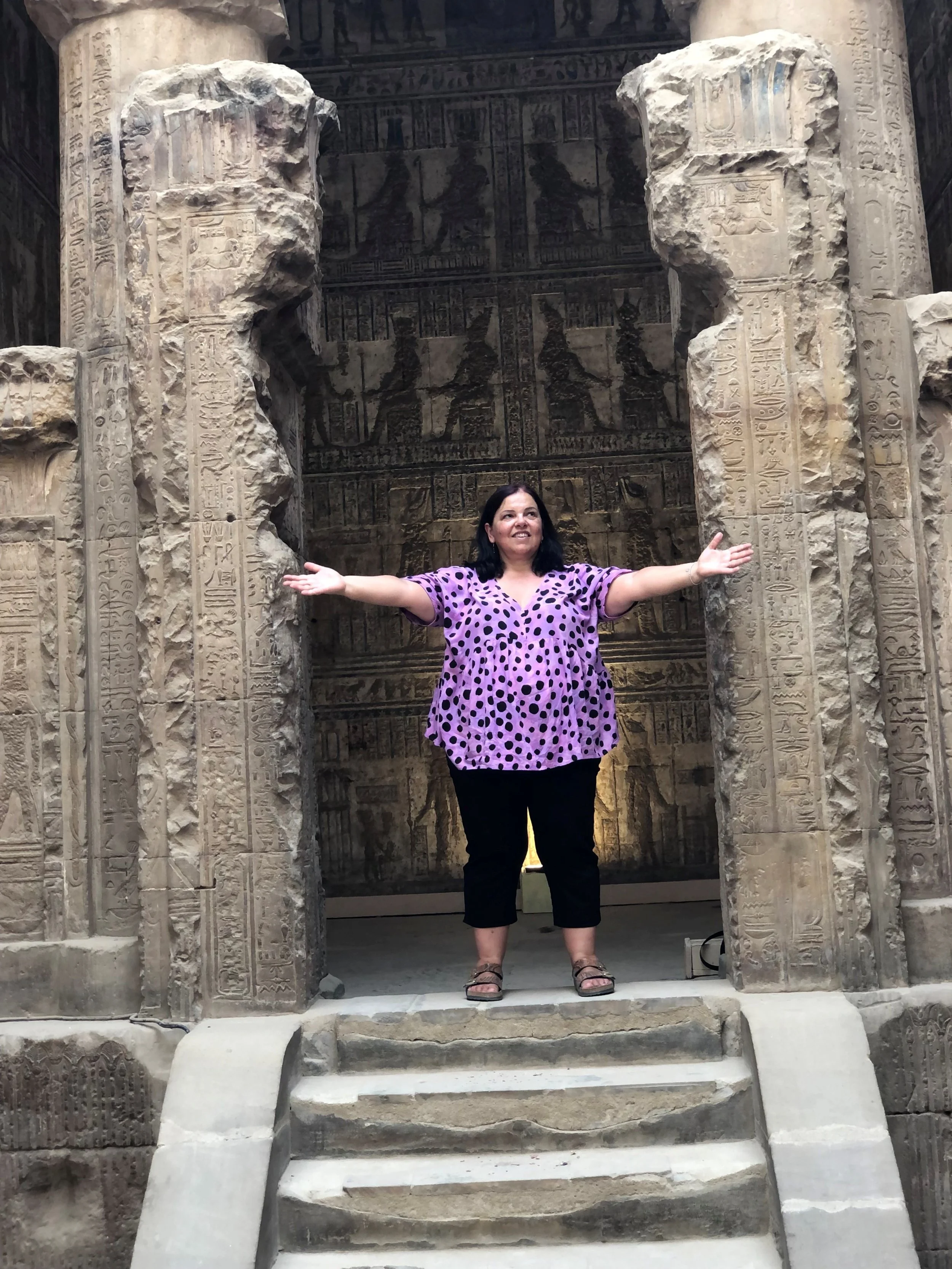 A woman standing at the entrance of an ancient Egyptian temple, with her arms outstretched and smiling. The entrance features tall, carved stone columns with Egyptian hieroglyphics and imagery, and a detailed wall relief depicting Egyptian gods and f
