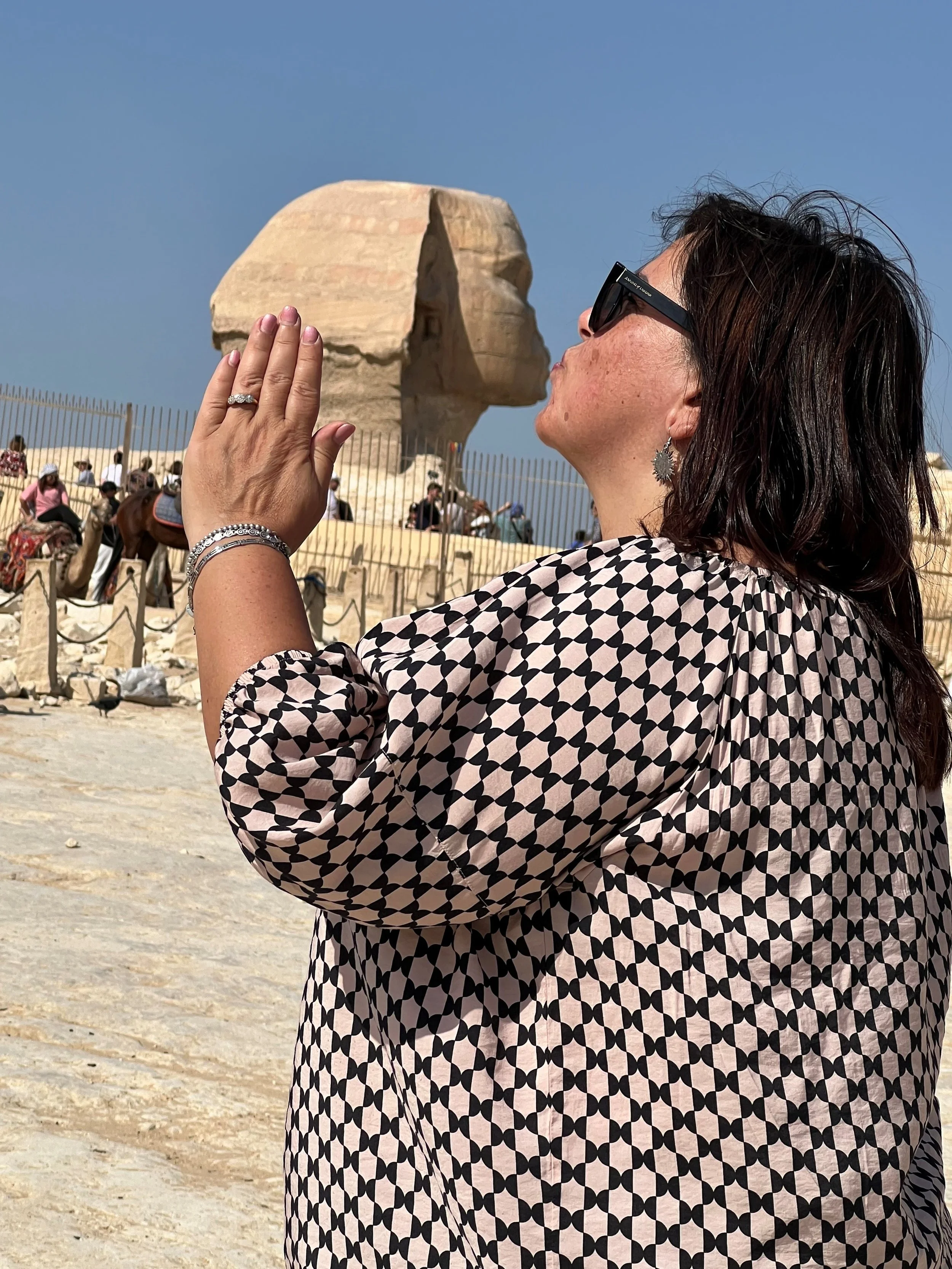 A woman with dark hair, sunglasses, and earrings is kissing the nose of the Sphinx statue at Giza, Egypt, during daylight with a clear blue sky.