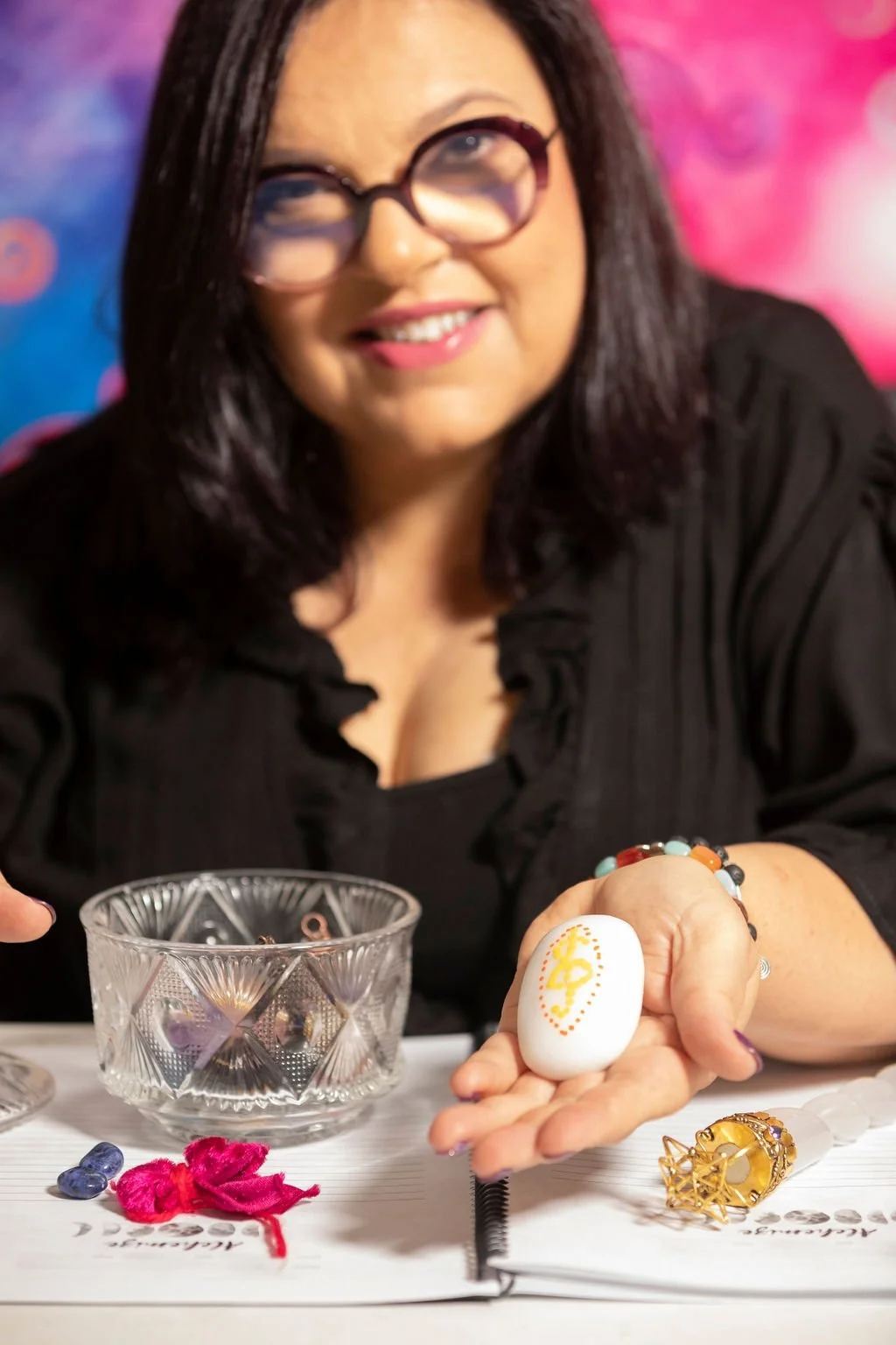 A smiling woman with glasses sitting at a table, holding a decorated stone with a yellow symbol, surrounded by jewelry and craft supplies.