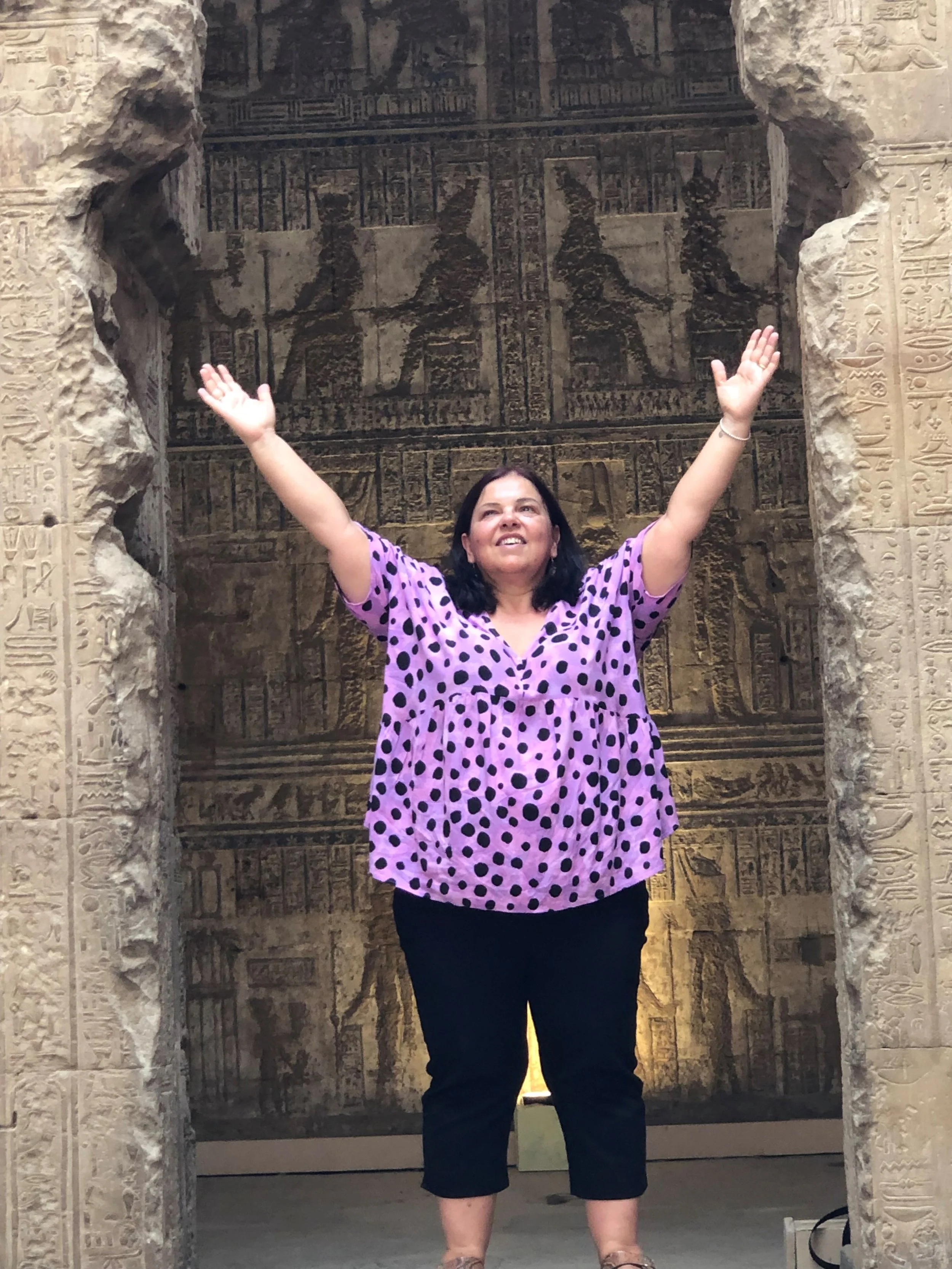 A woman standing with arms raised in front of ancient Egyptian hieroglyphic walls, framed by stone columns with hieroglyphs.