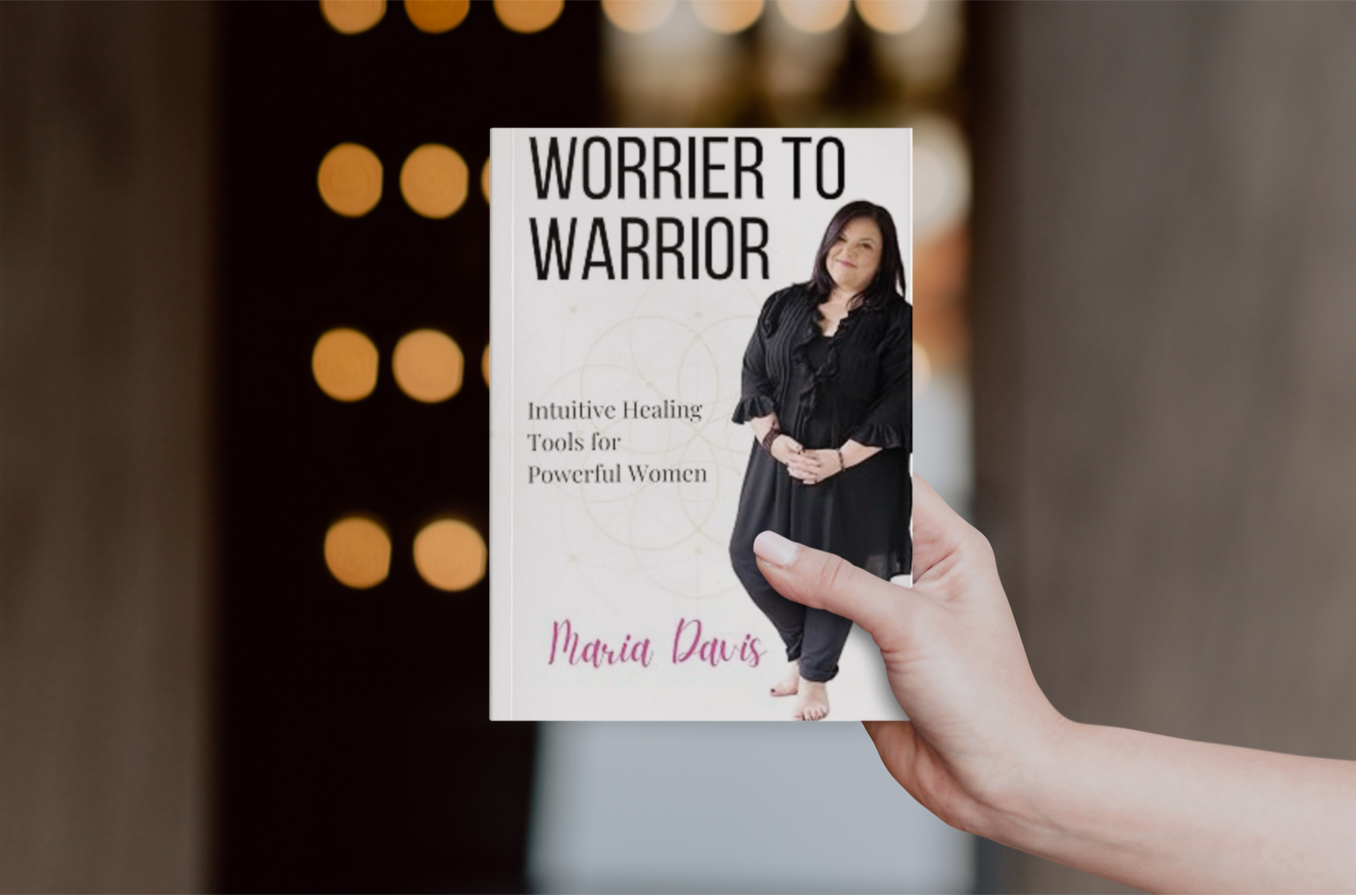 A person holding a book titled "Worrier to Warrior" by Maria Davis. The book cover features a photo of a woman with dark hair, wearing a black outfit, and smiling. The background of the photo has circular light spots.