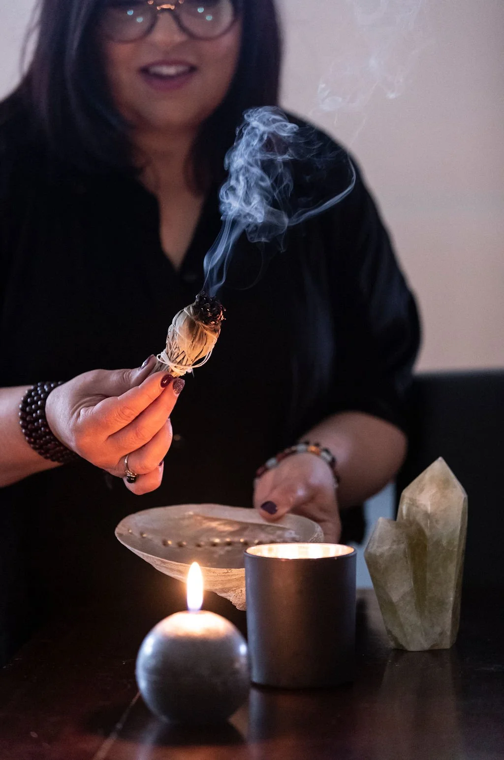 A woman with glasses and dark hair smiling and holding a small sage smudge stick with smoke rising from it, surrounded by lit candles and crystals on a dark table.