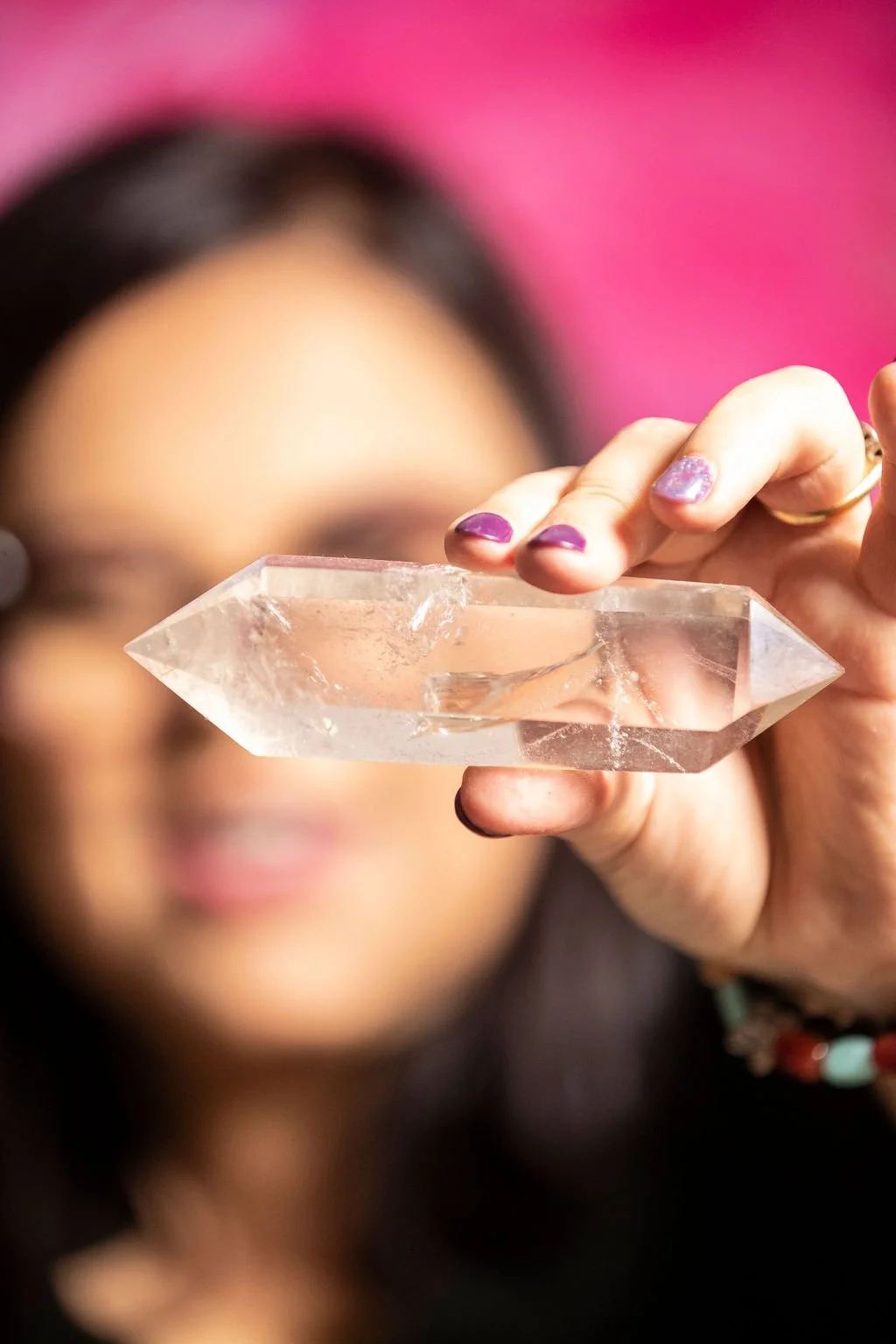 Person holding a clear quartz crystal pointed towards the camera with a blurred face and pink background.
