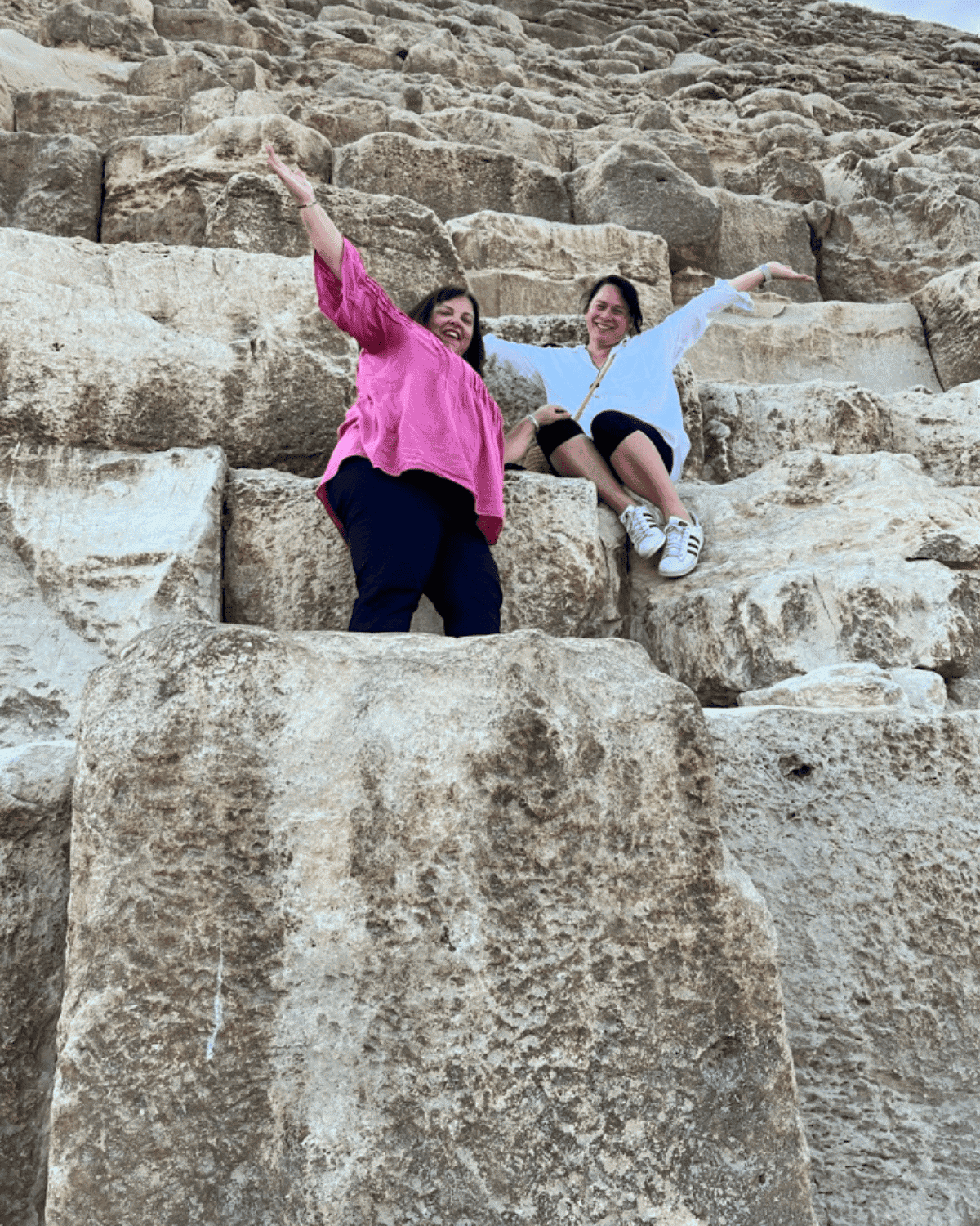Two women sitting on large rocks in a desert landscape, smiling and raising their arms in celebration.