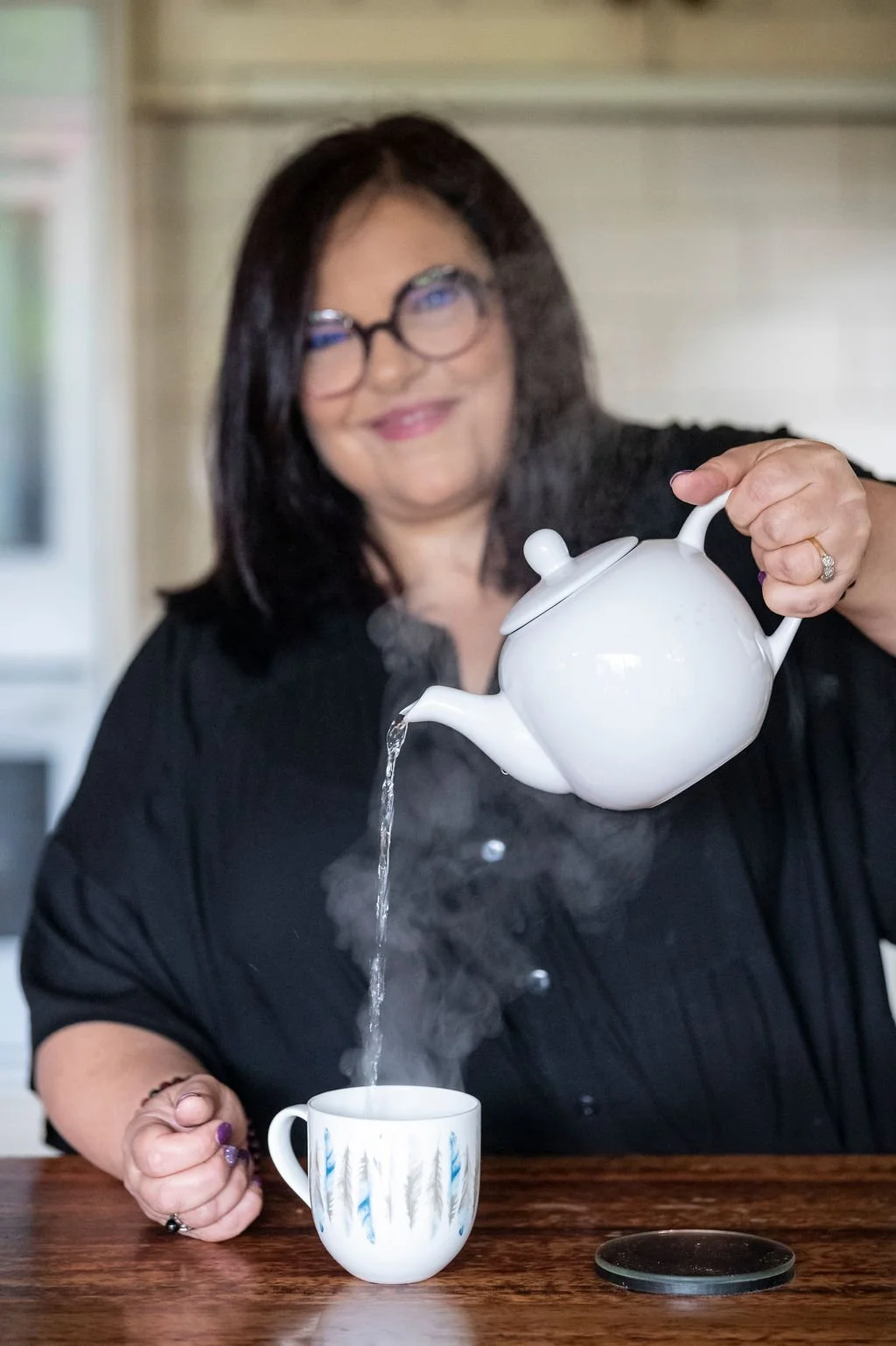 A woman with dark hair and glasses pouring hot water into a mug, with steam rising from it.