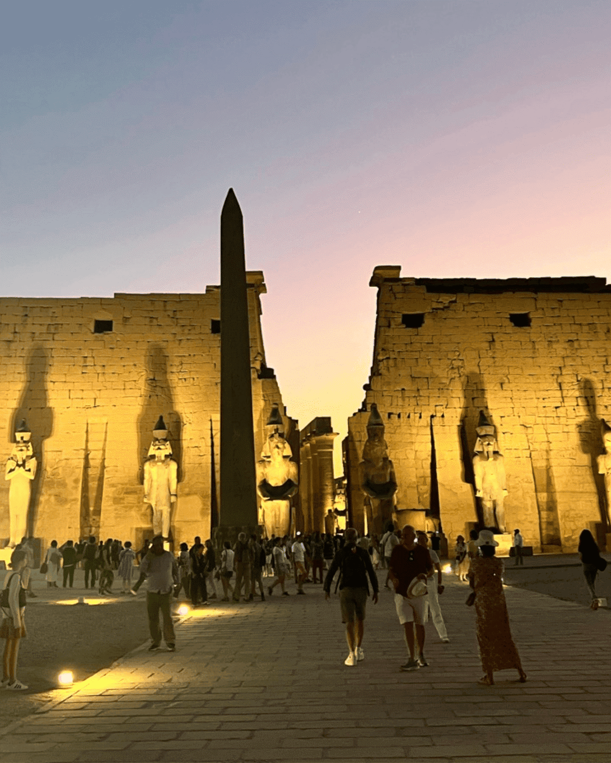People walking at sunset in front of the Luxor Temple in Egypt, with large statues of Egyptian gods and an obelisk.