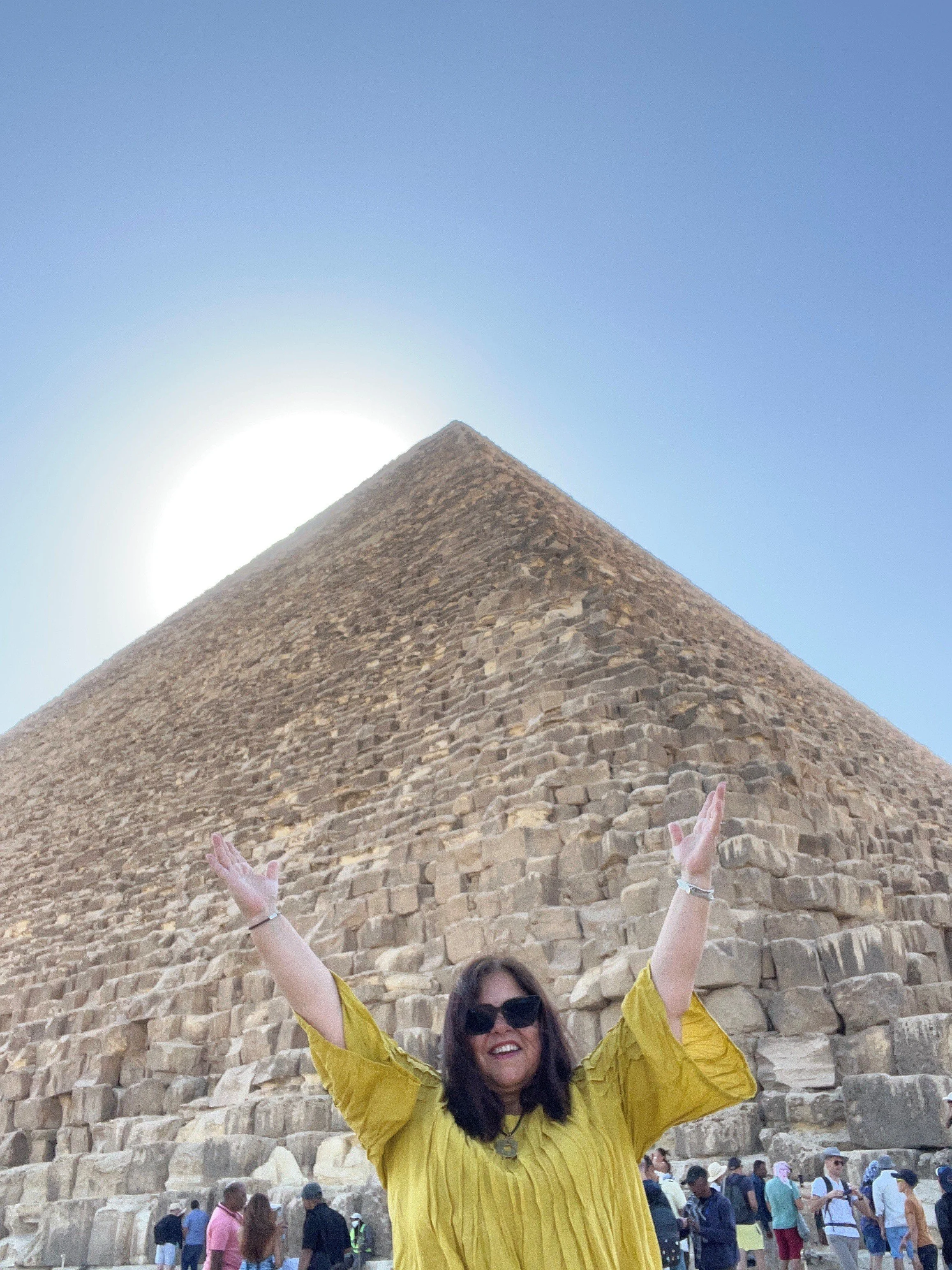 A woman wearing sunglasses and a yellow shirt with her arms raised, standing in front of the Great Pyramid of Giza surrounded by tourists.