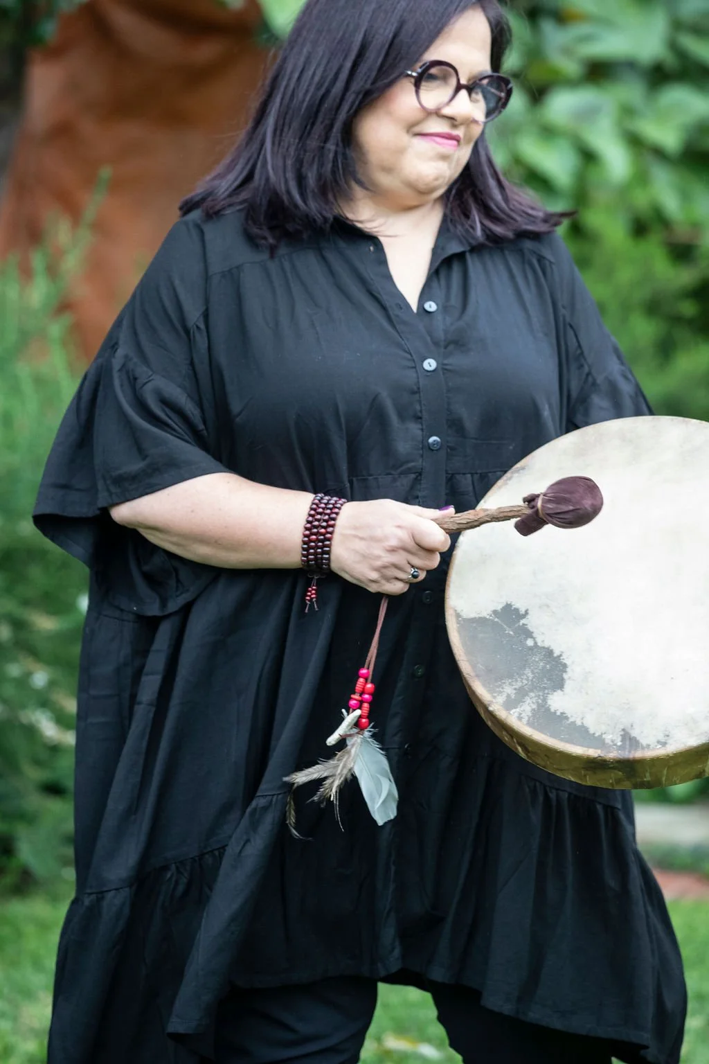 A woman with dark hair and glasses wearing a black dress, holding a wooden drum with a mallet. She is outdoors with green foliage in the background.