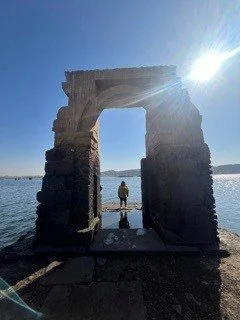 A stone arch structure on a beach with two people standing underneath it, water and sky in the background, and sunlight shining brightly from the top right.