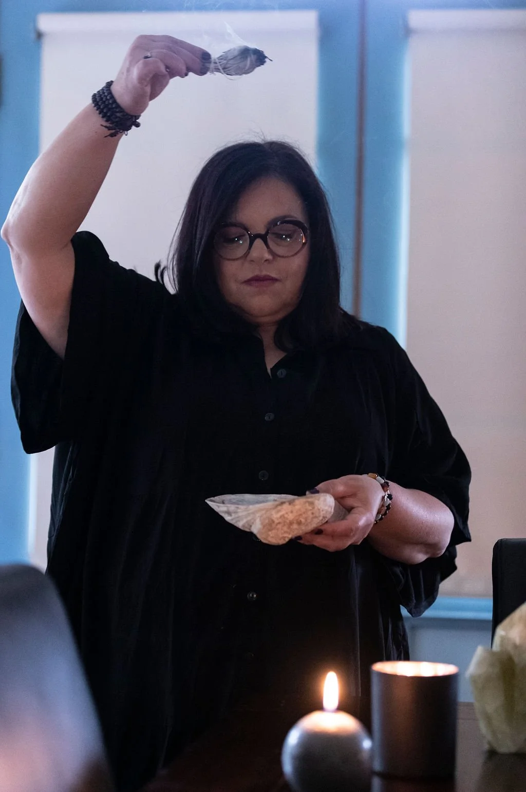 A woman with black hair and glasses holding a seashell in one hand and a small bag in the other, with a candle and another object on a table in the foreground.
