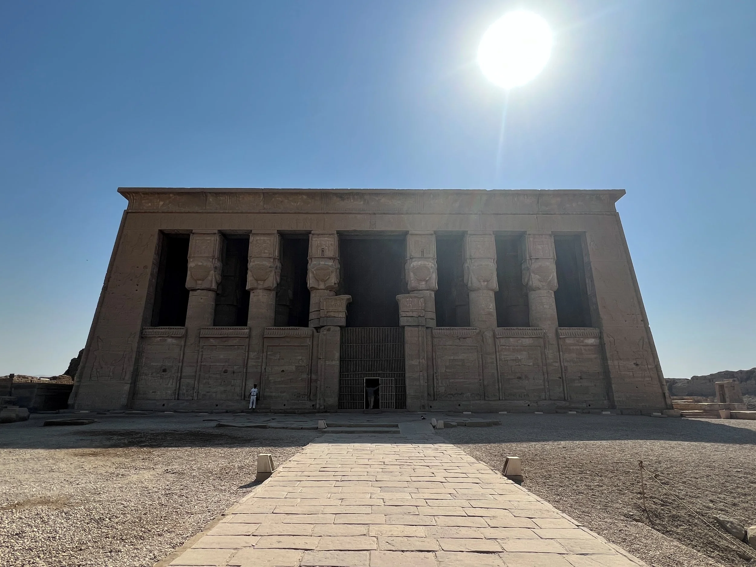 The front view of an ancient Egyptian temple with a large stone structure, columns, and a pathway leading up to it, under a bright sun in a clear blue sky.