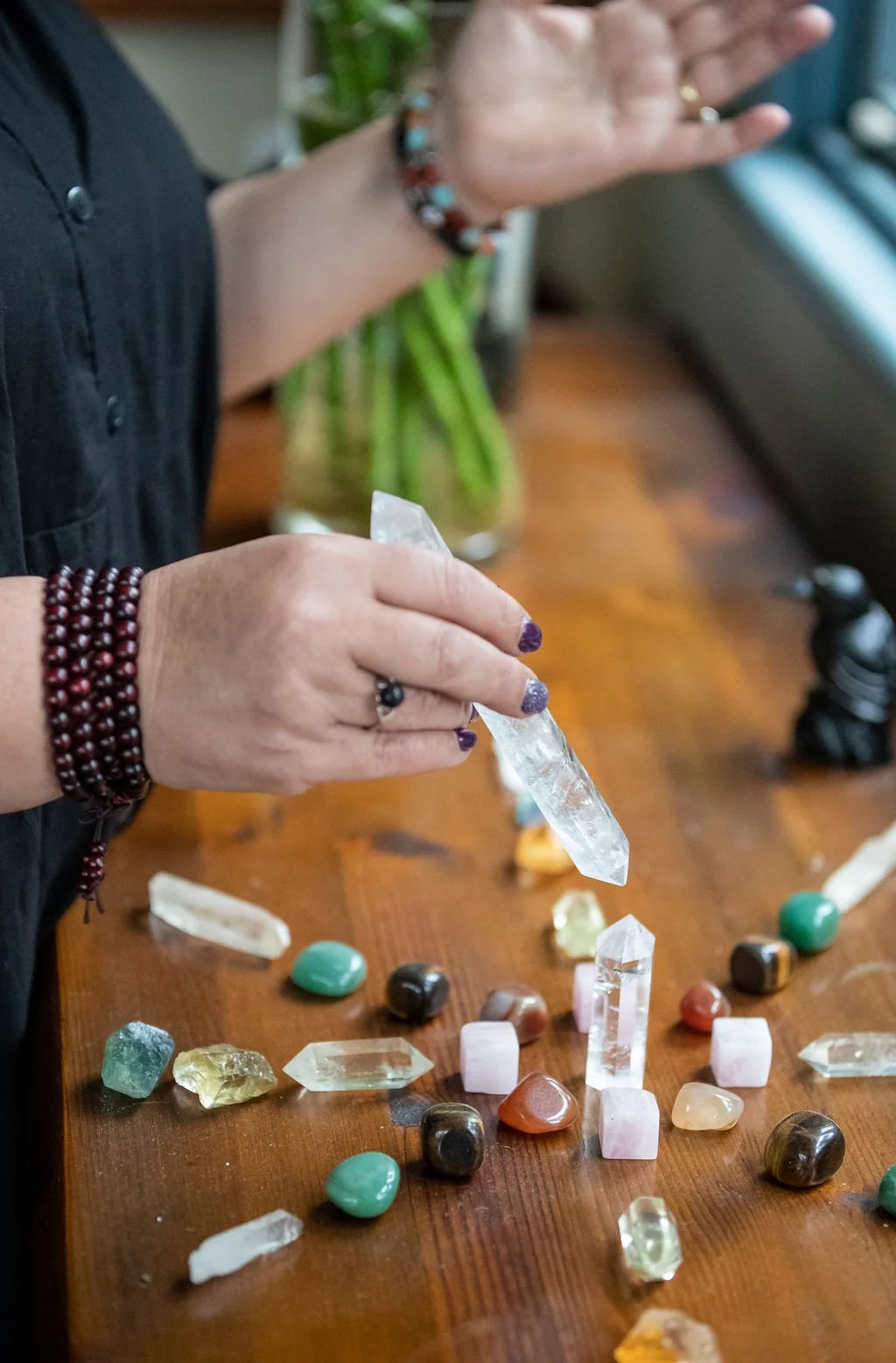 Person handling a large clear quartz crystal over a table with various smaller polished and raw gemstones, with a potted plant in the background.