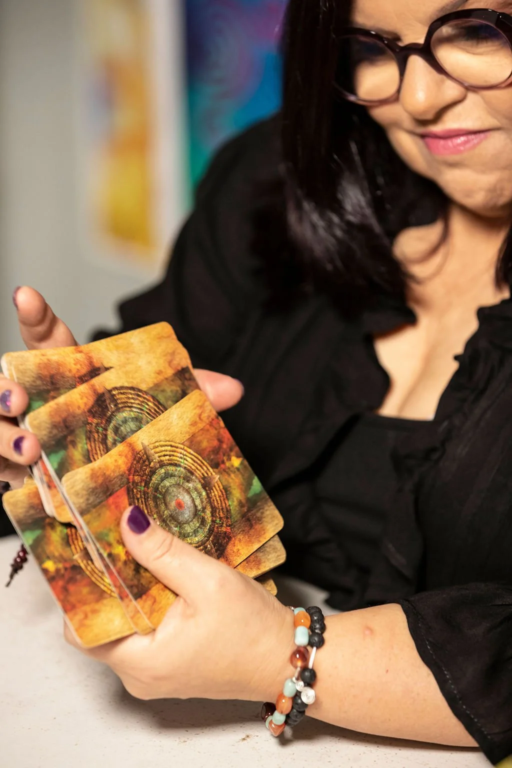 A woman with glasses holds a spread of tarot cards with an abstract, colorful design. She is smiling slightly and wearing a black top and beaded bracelets.